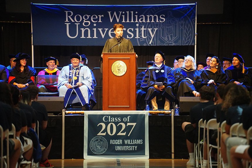 An RWU student stands at the podium with other speakers sitting on the stage on either side of them