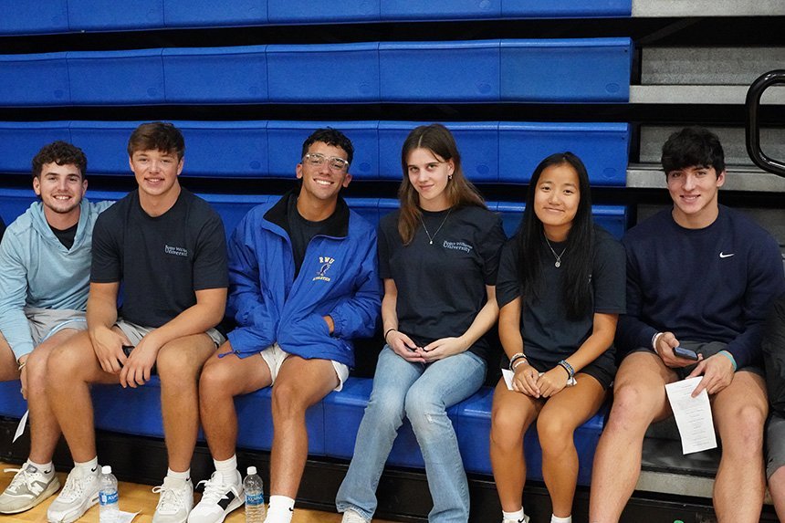 Six students wearing navy blue RWU t-shirts sit on bleachers and smile at the camera