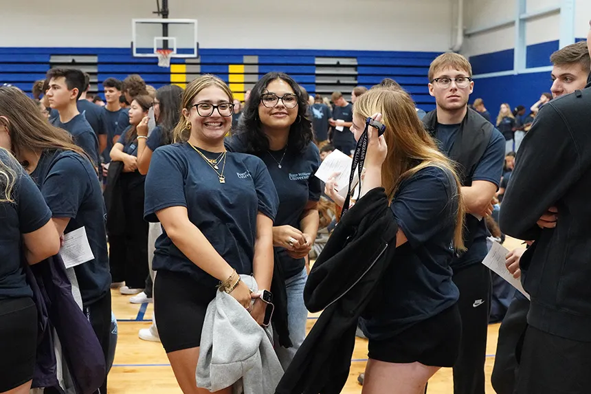 Two students standing and smiling at the camera in a gymnasium, surrounded by other students, all wearing blue t-shirts