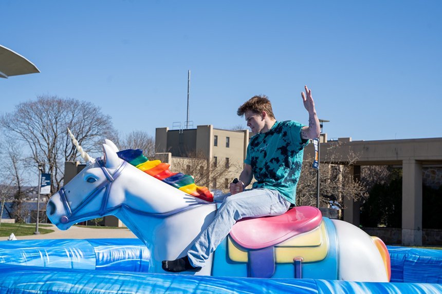 A student rides a mechanical unicorn on the green in front of the library as part of Queer Fest.