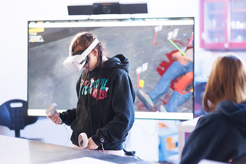 A student wearing a VR headset in front of a big screen showing a virtual crime scene.