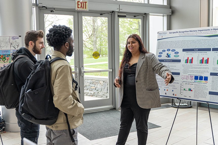A student presenting standing in front of her research poster talking to two other students at SASH.