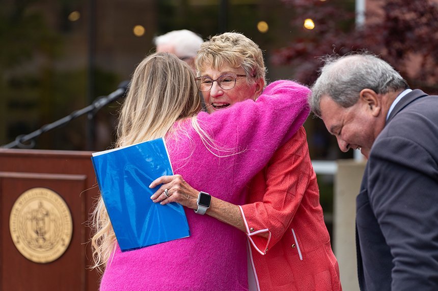 Joyce Cummings and Heidi Maes hugging as President Ioannis Miaoulis stands by