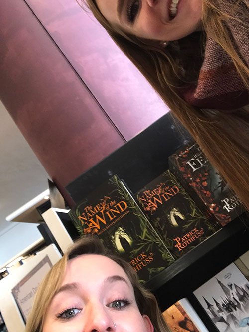 Two people take a selfie in front of The Name of the Wind by Patrick Rothfuss