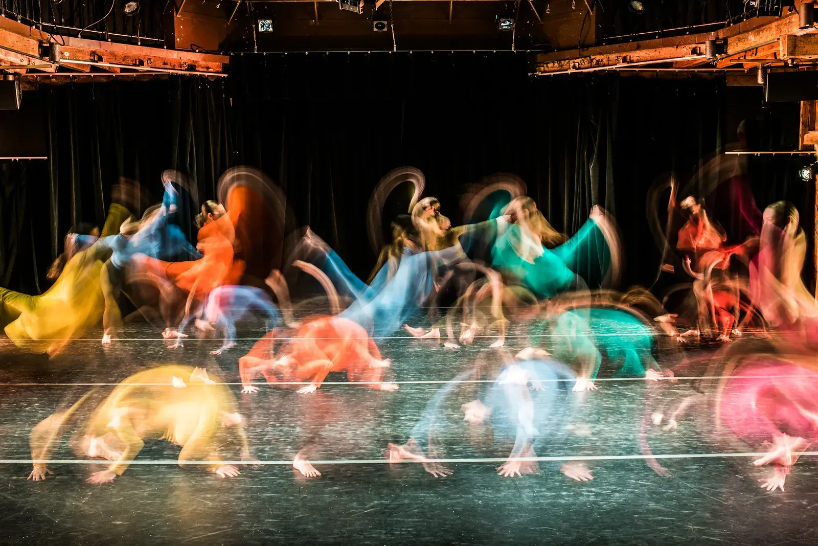 Dancers in colorful costumes perform on stage, with motion blur capturing energy and movement against a dark backdrop.