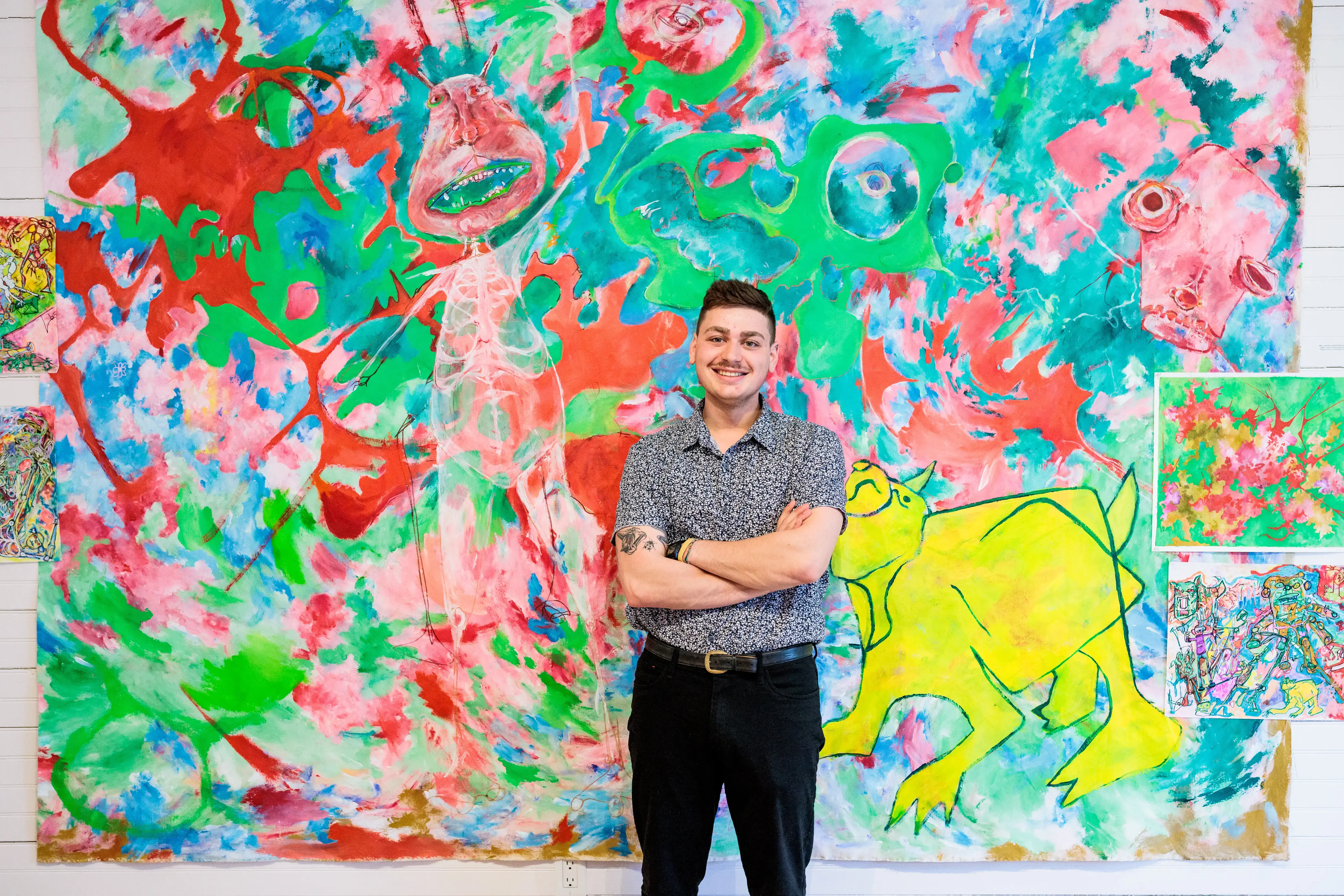 A smiling person stands proudly in front of a bold, colorful painting with surreal figures, posing with their own artwork.