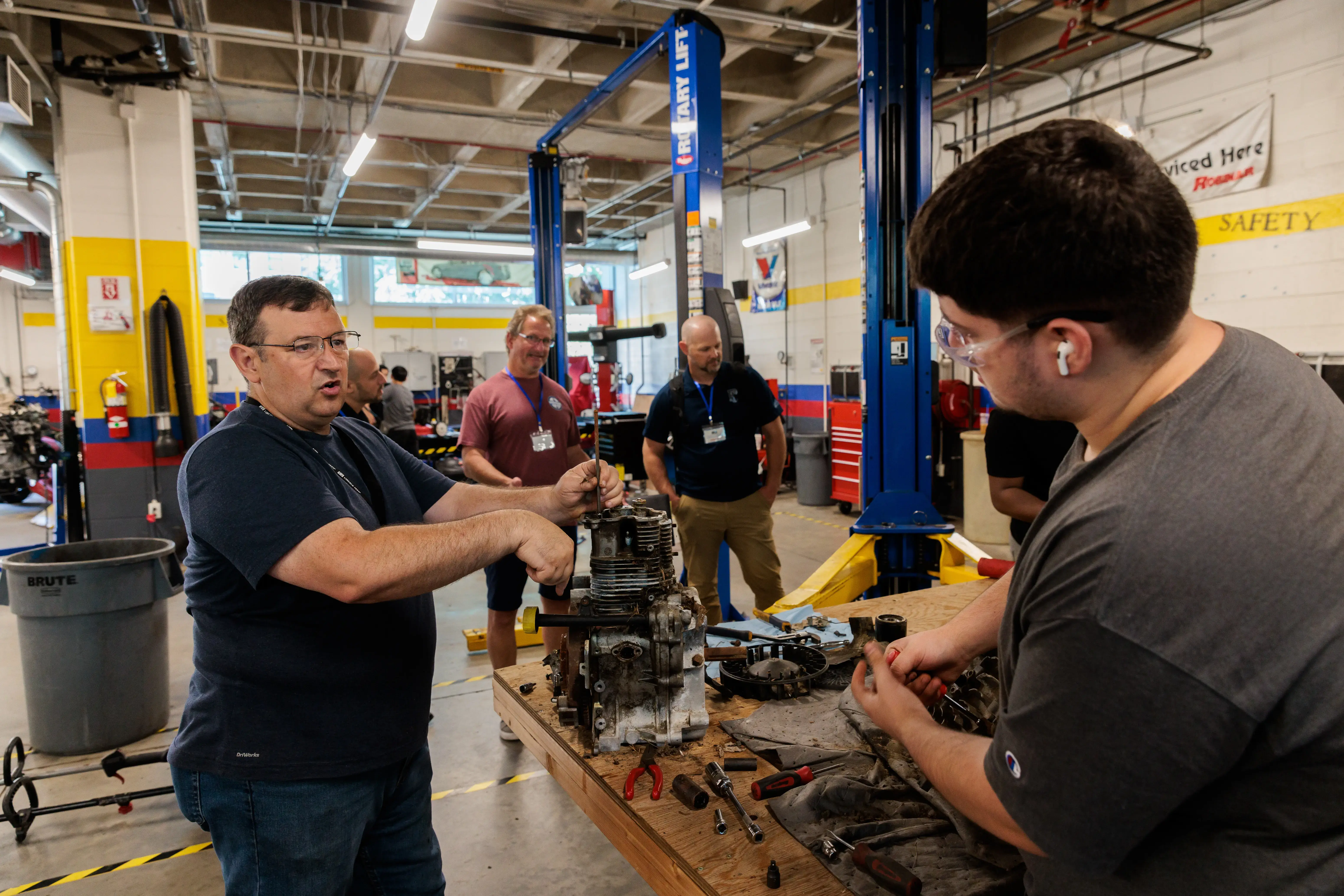 An instructor and student work on a disassembled engine at a workshop table, while others observe in an auto tech classroom.