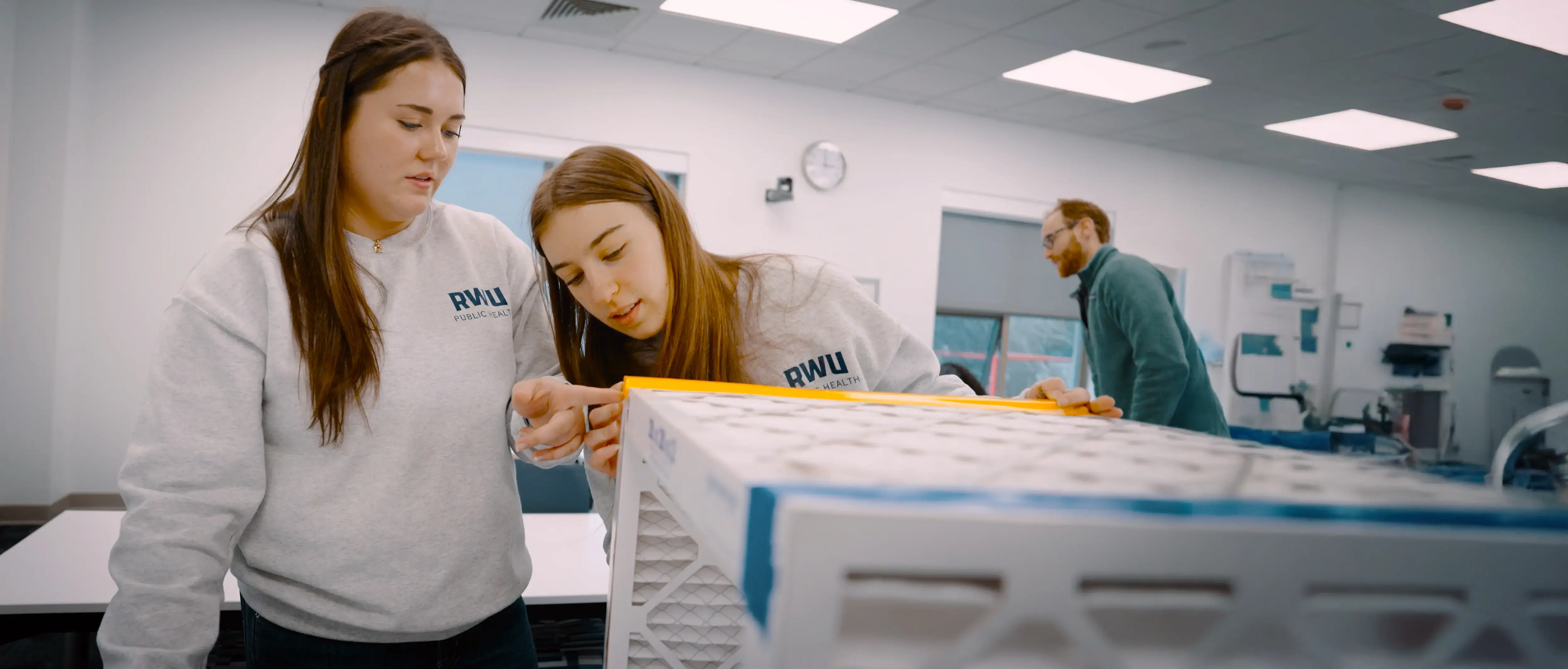 Two students in RWU Public Health shirts measure a filter unit in a classroom while a man works in the background.
