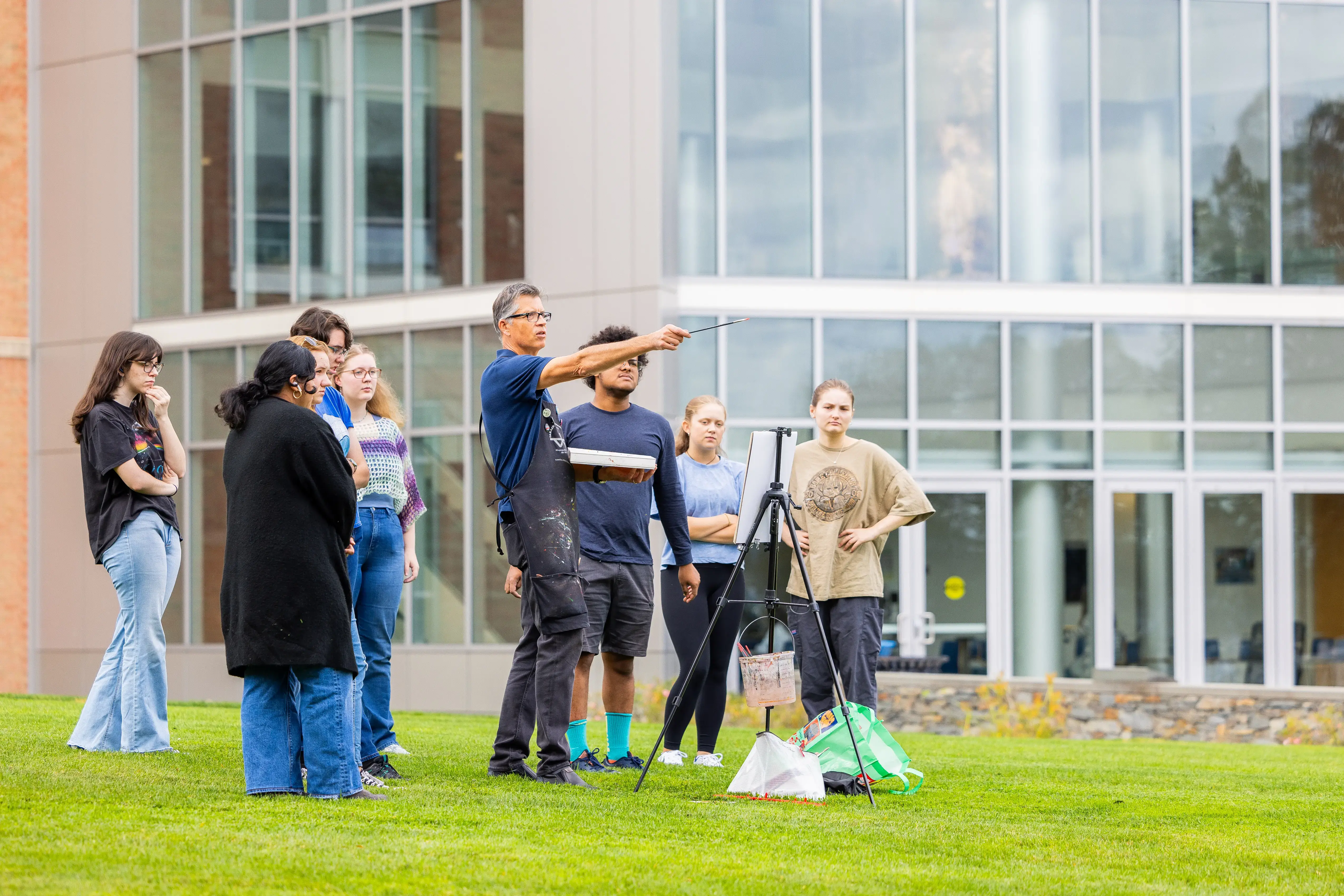Students gather around a professor as he give a painting lesson while outdoors.