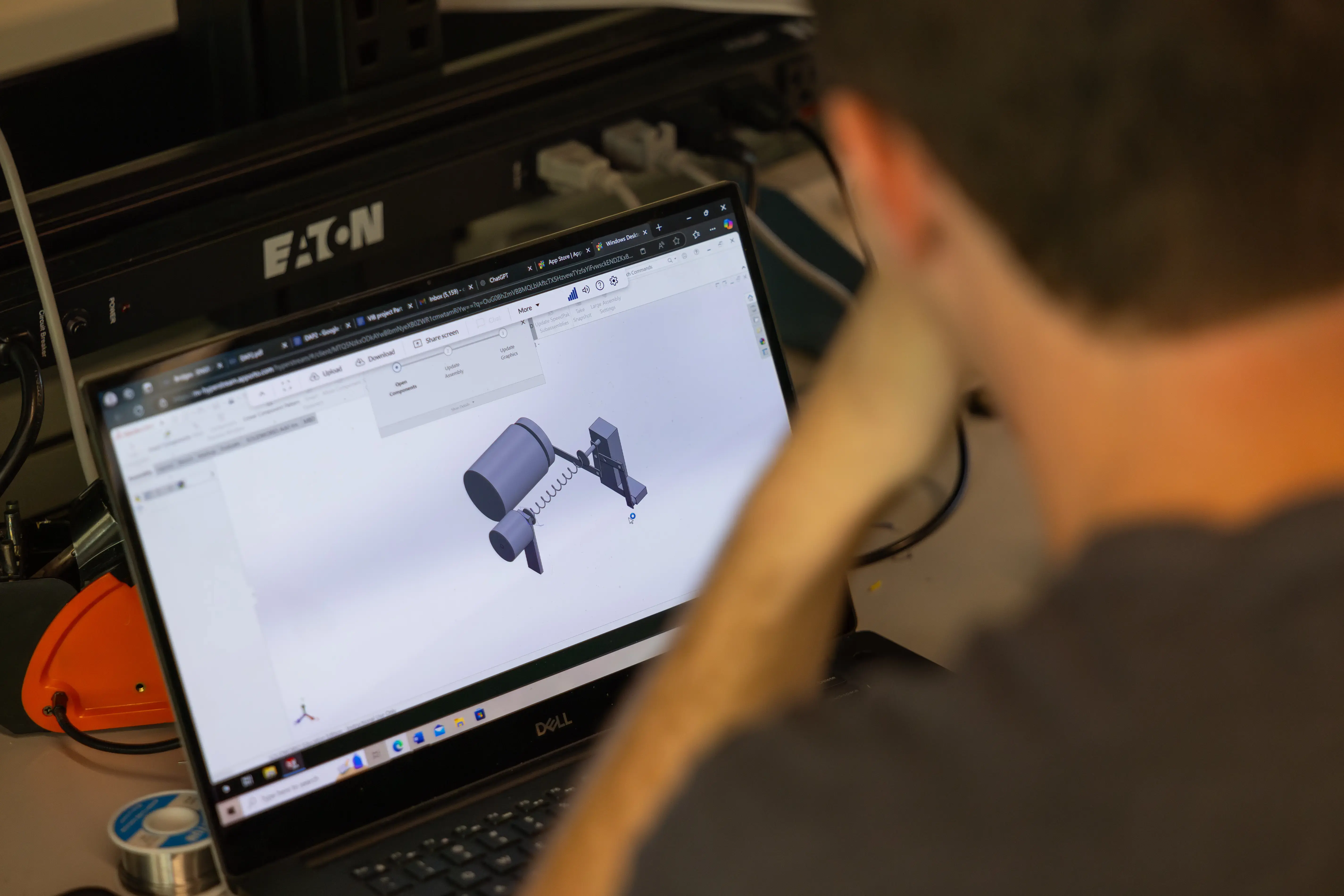 A person examines a mechanical 3D CAD model on a Dell laptop at a workstation with tools and electronics nearby.