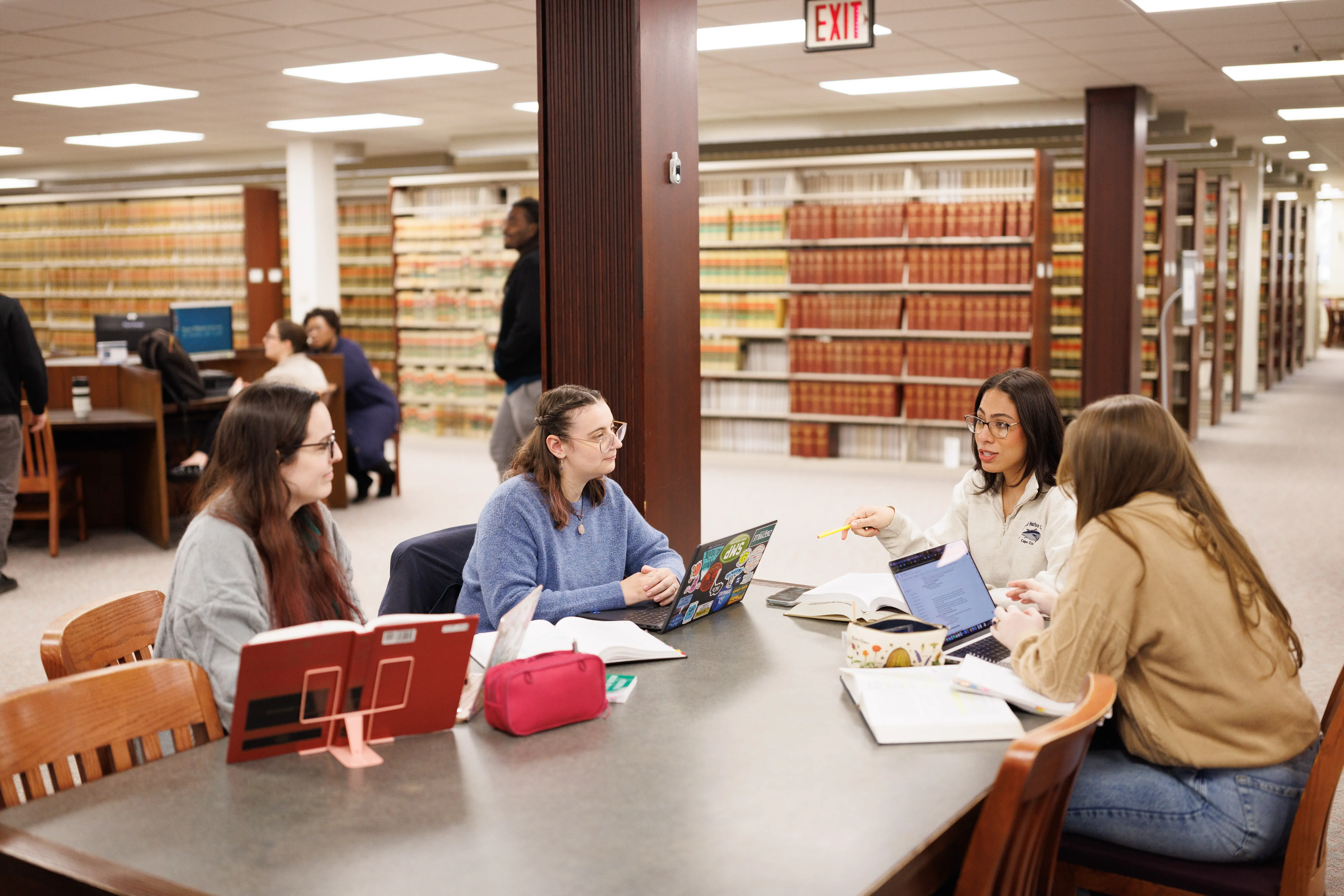 Four students study together at a library table with laptops and books, surrounded by shelves of legal volumes.