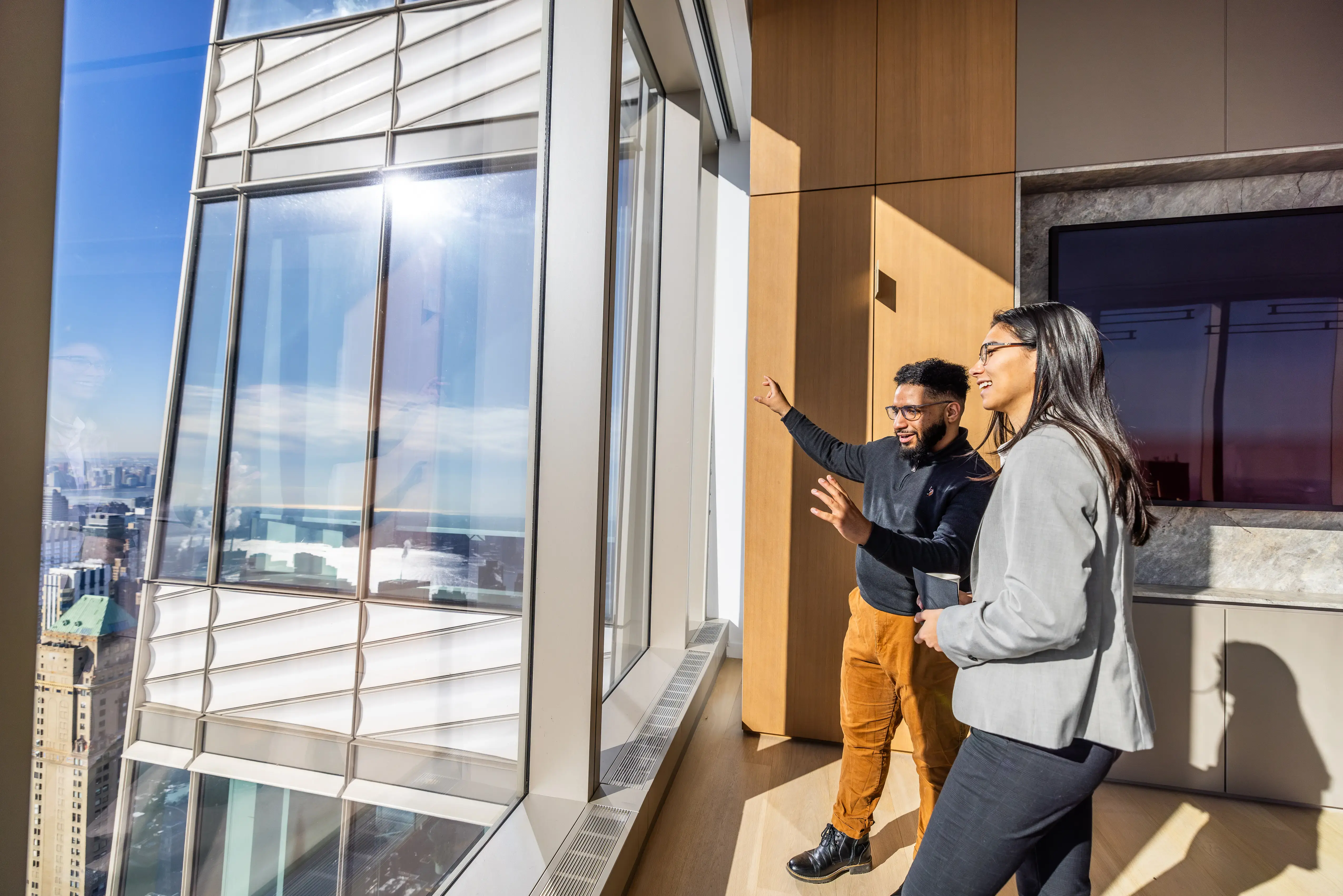 Two professionals stand by a large window in a modern office, viewing the city skyline as one gestures and the other smiles.