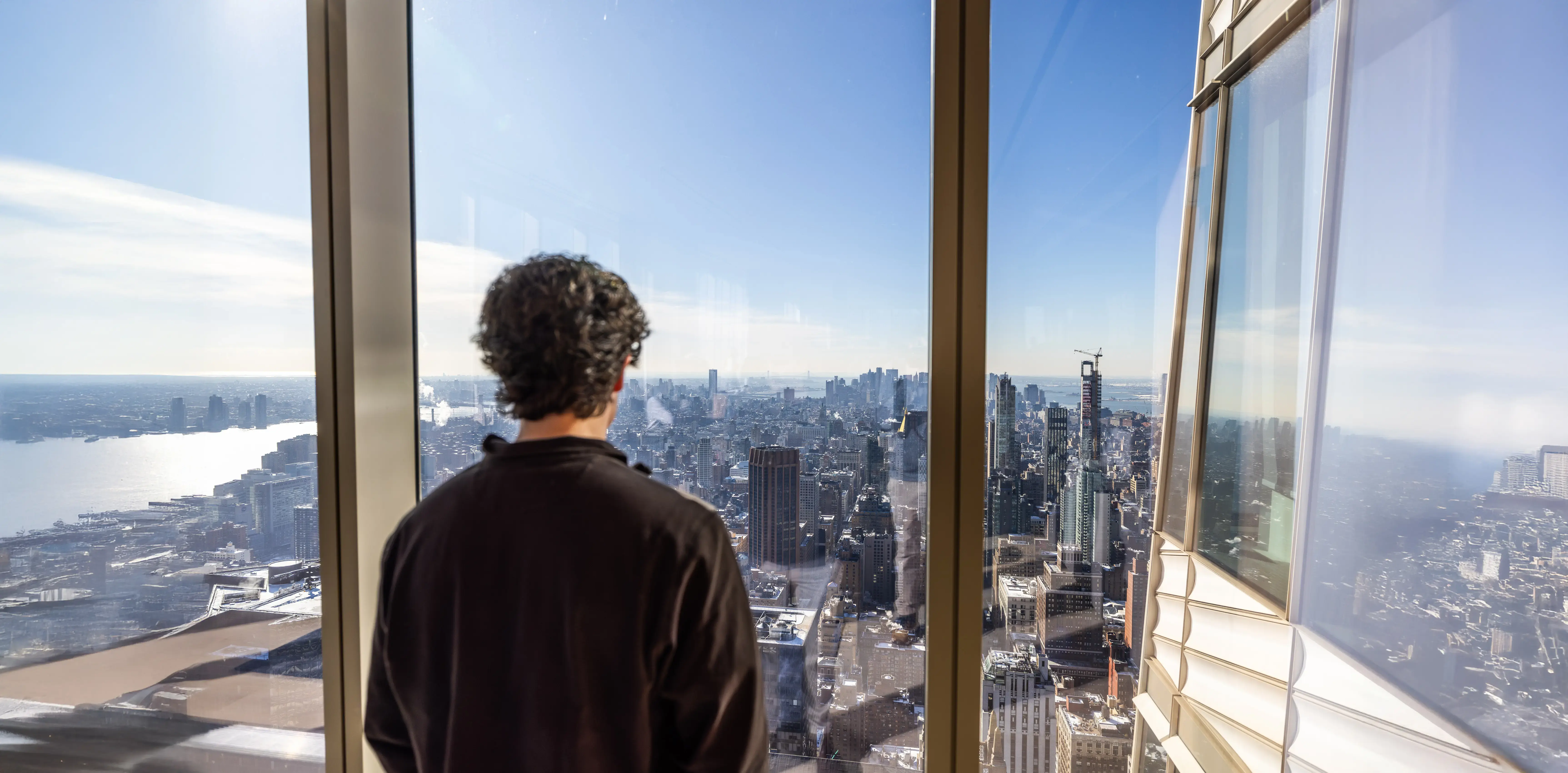 Urban Studies student looks out tall window at sunny city skyline and river, with building reflections visible on the glass.