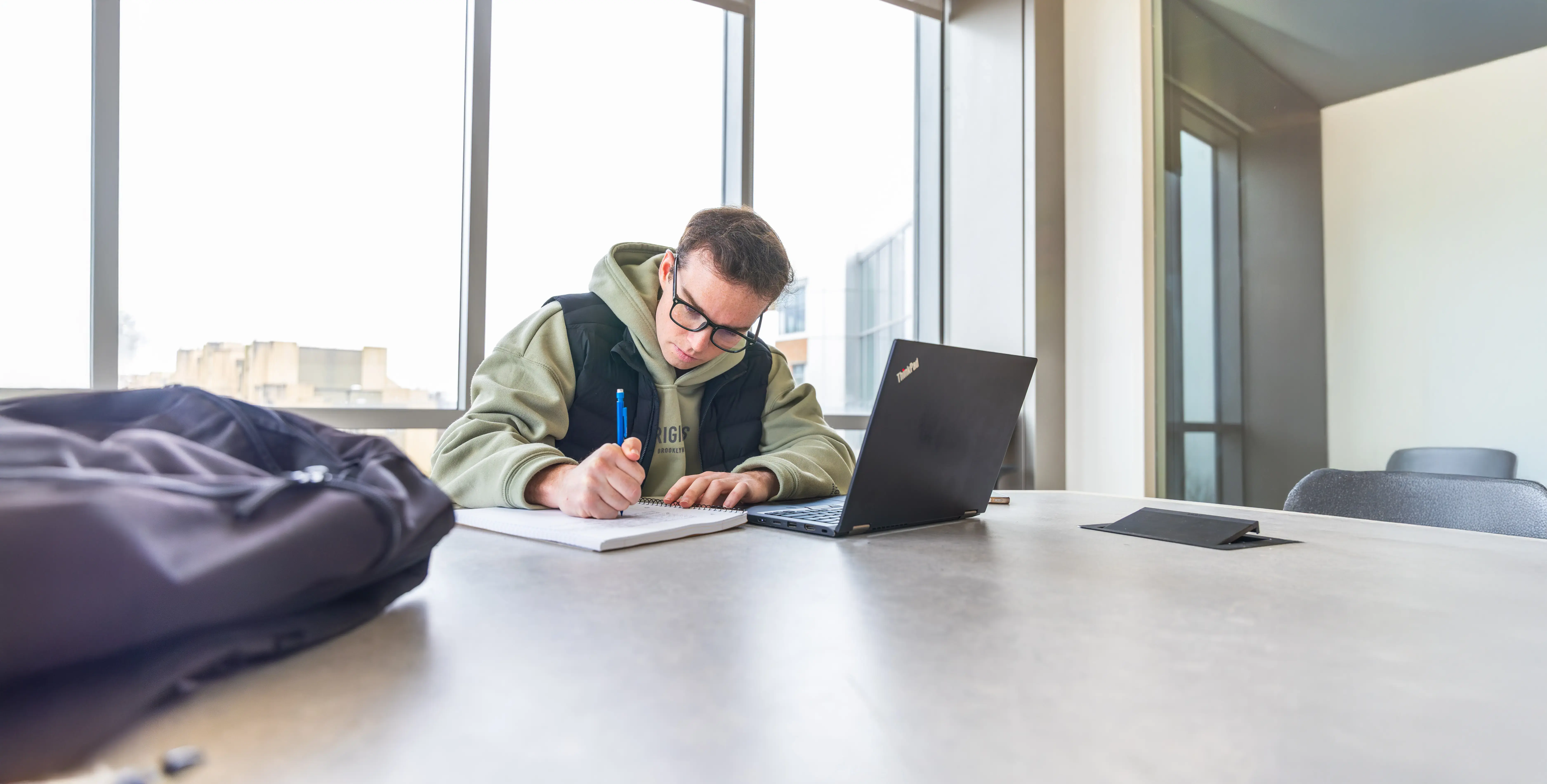 Student wearing glasses writes notes beside laptop in bright study room with large windows and empty chairs.