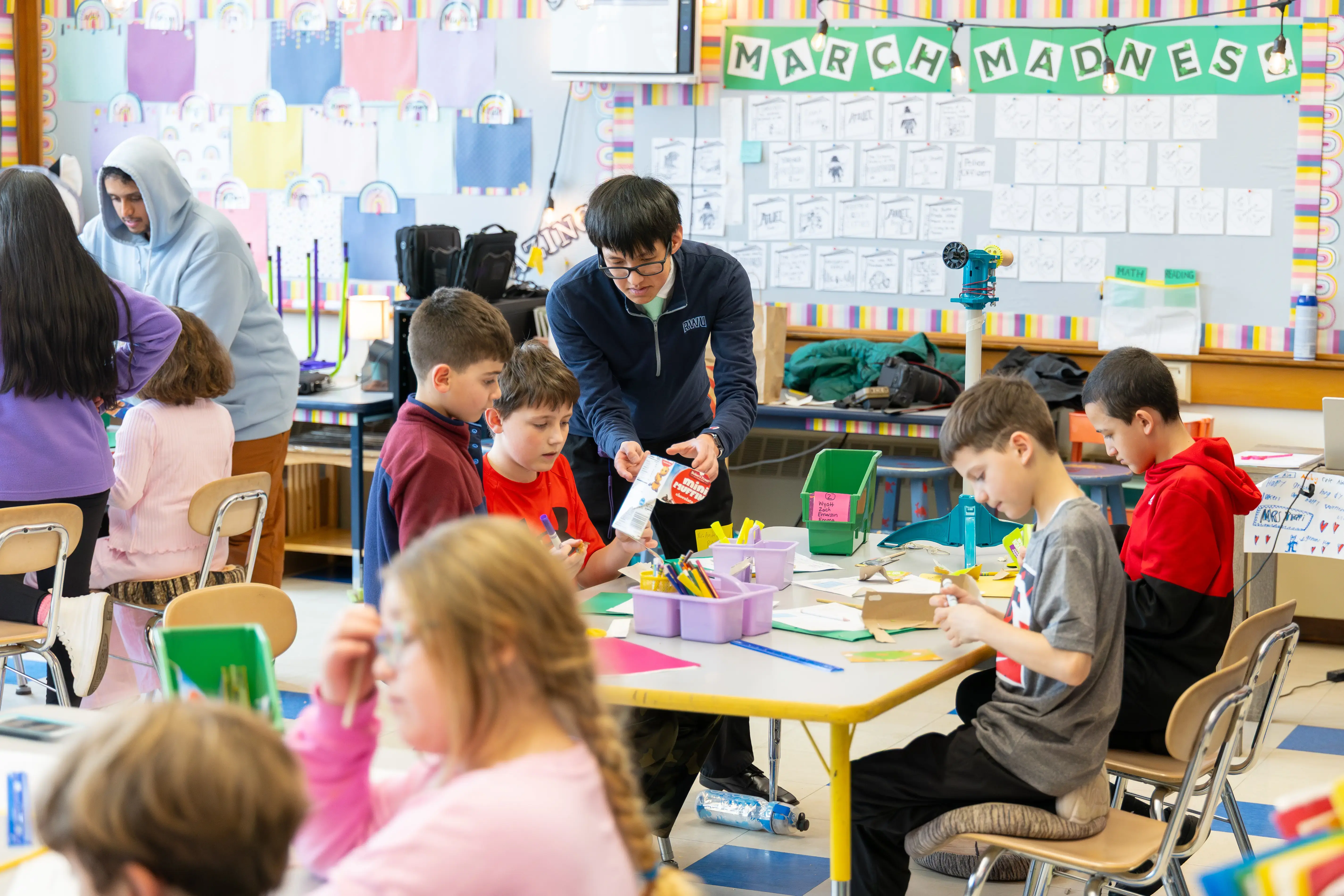 Kids do crafts as an adult helps two boys under a “March Madness” bulletin board in a colorful classroom.
