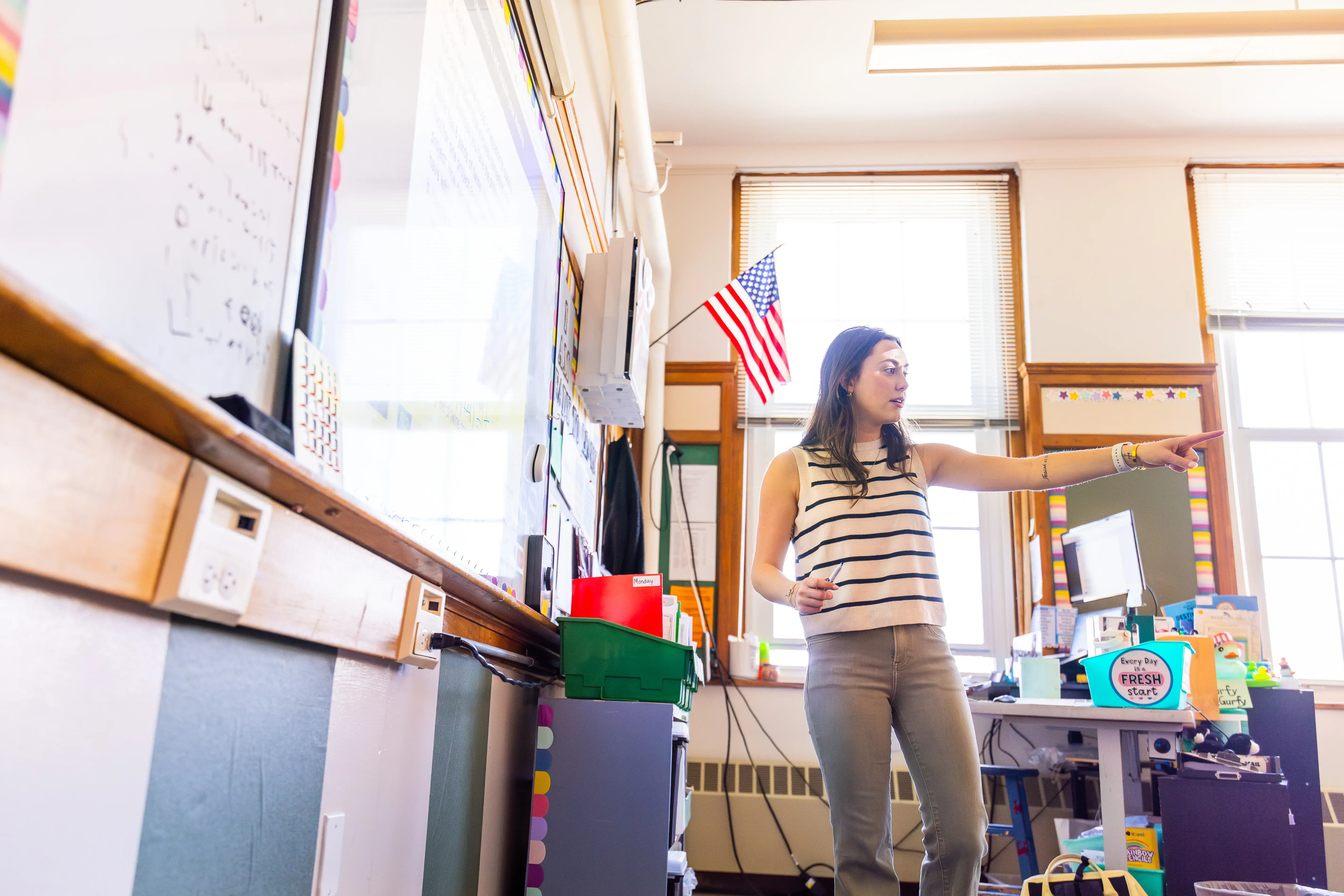 A teacher stands at the front of a middle school classroom, pointing while speaking, and classroom supplies visible.