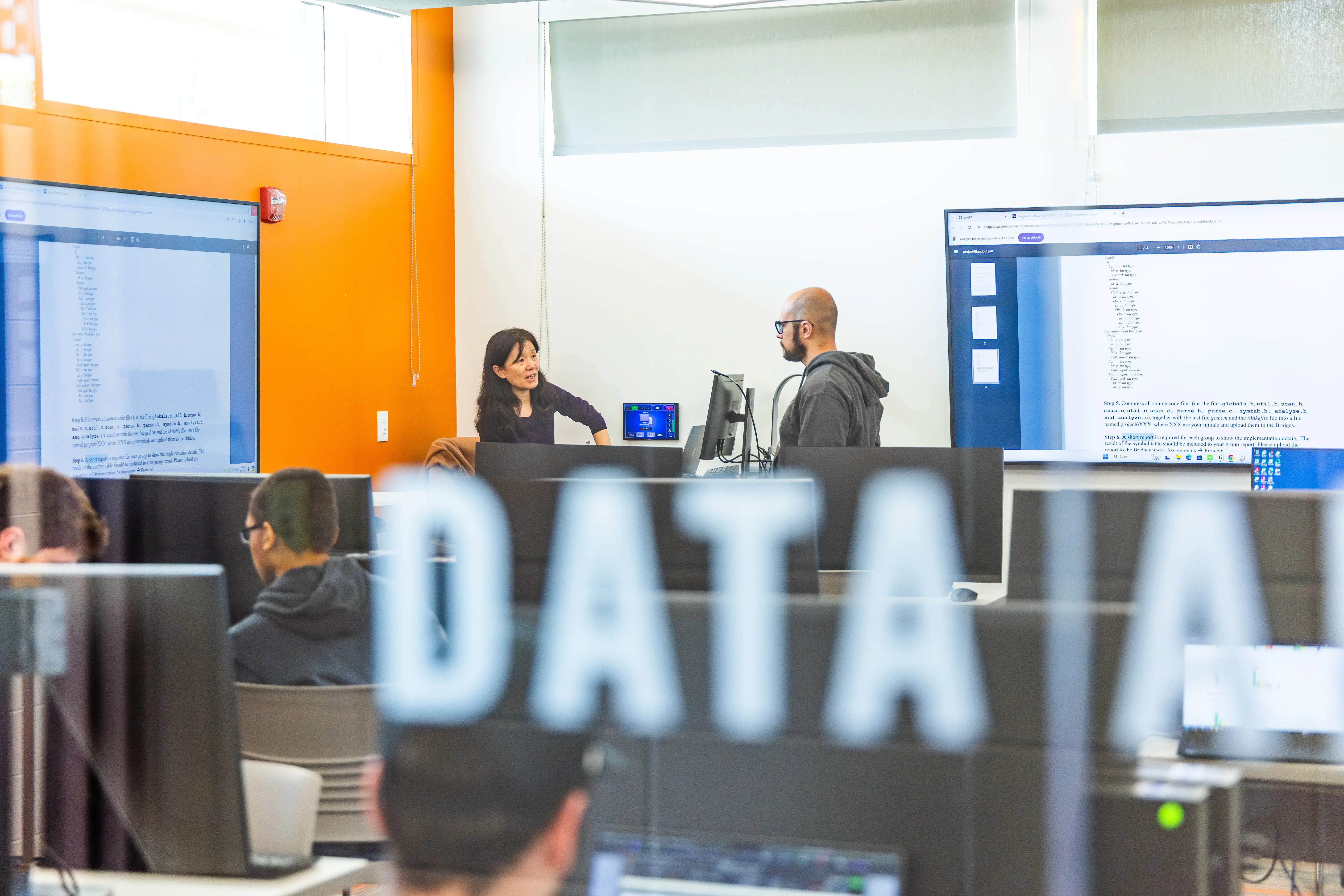 Students work at computers behind a glass wall labeled “DATA” while instructors lead a coding lesson in a bright classroom.