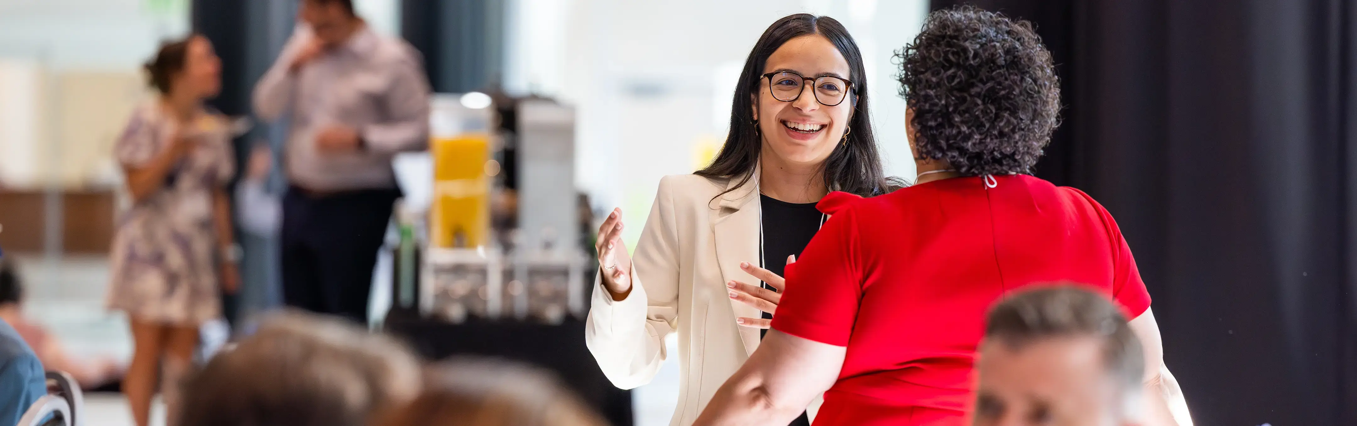 A smiling woman in a white blazer chats with another in red at a lively event near a table with juice dispensers.