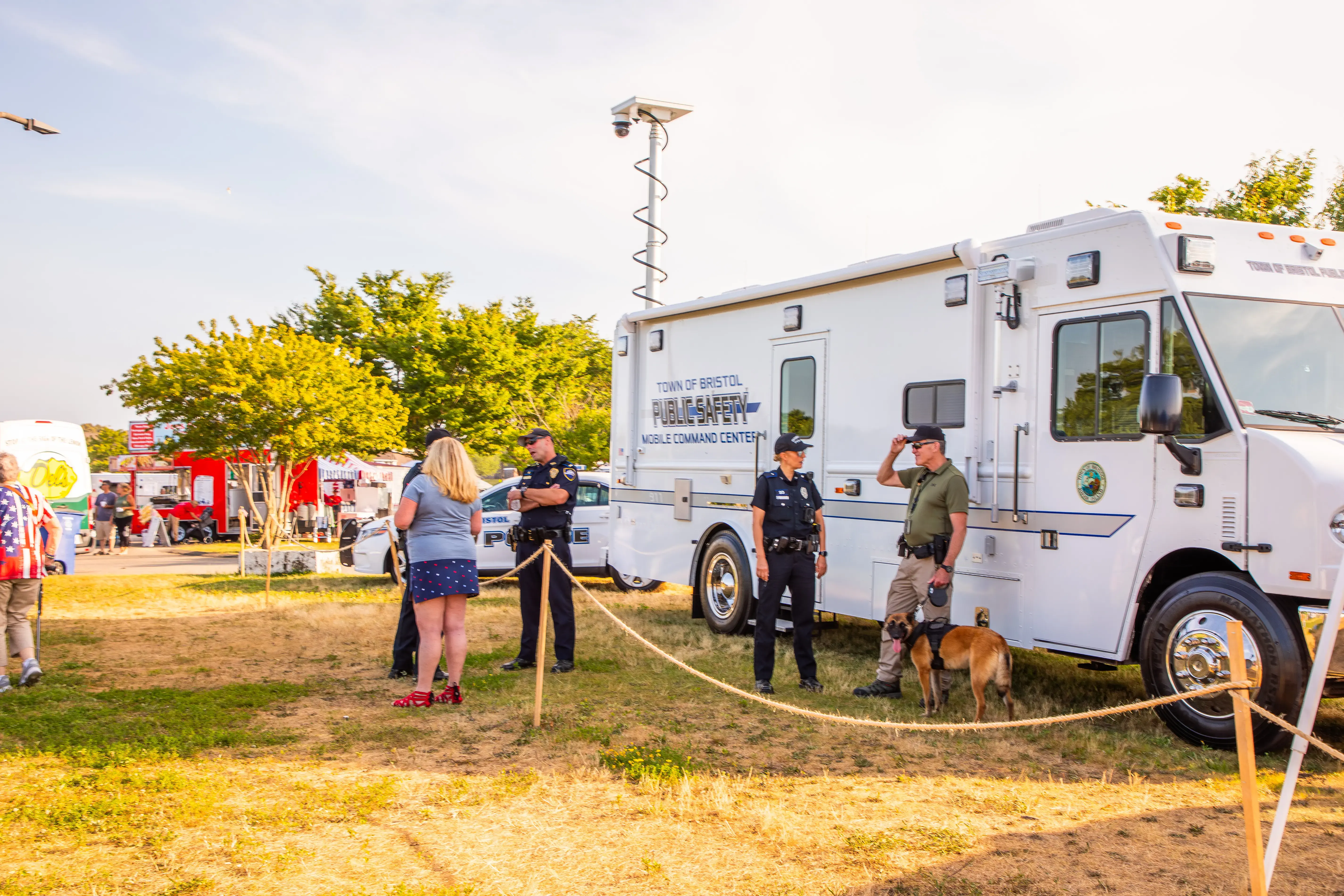 Public safety officers stand near a Mobile Command Center at an outdoor community event with a dog, tents, and vehicles.