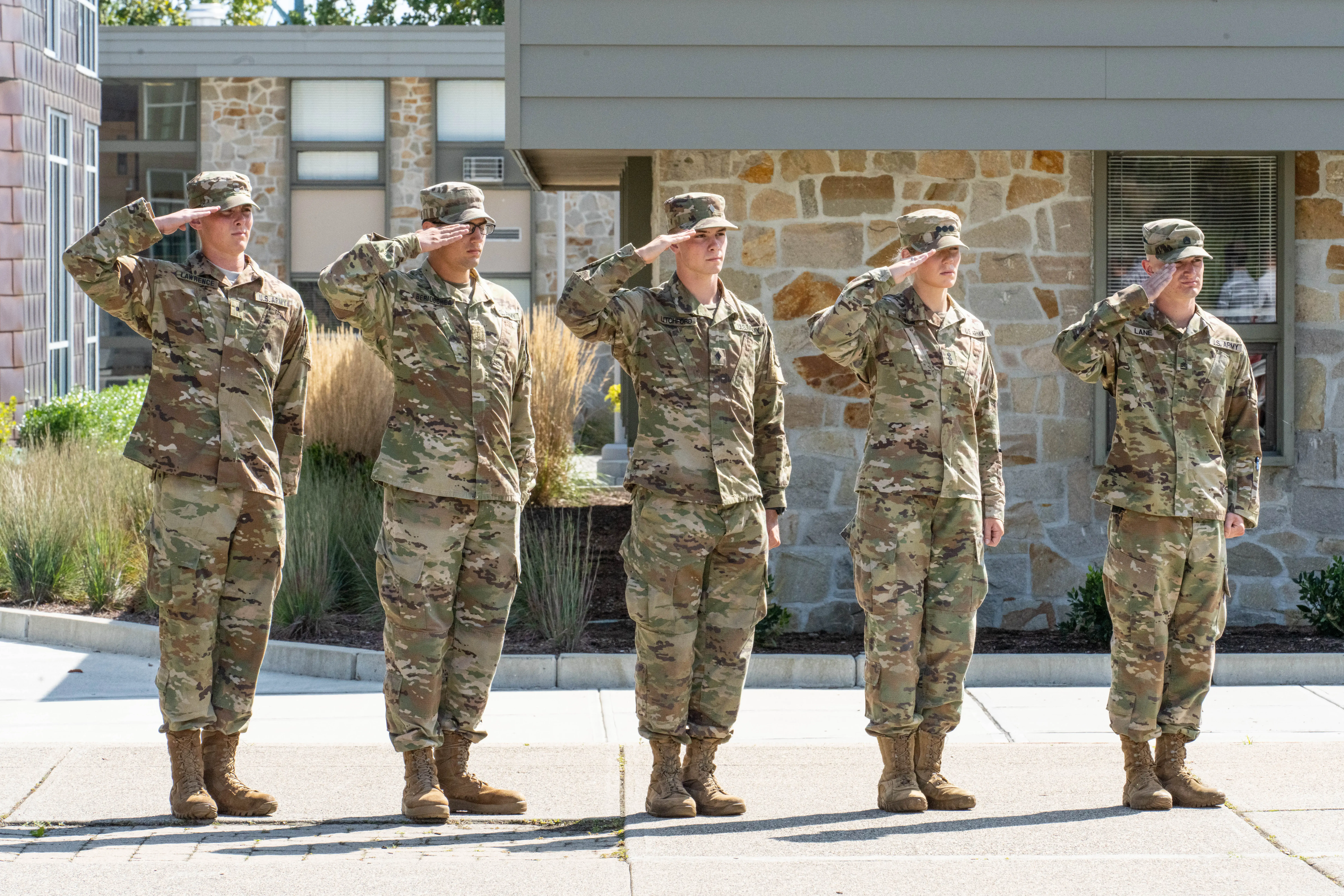 Five people in military uniforms salute in front of a stone building during a formal outdoor ceremony.