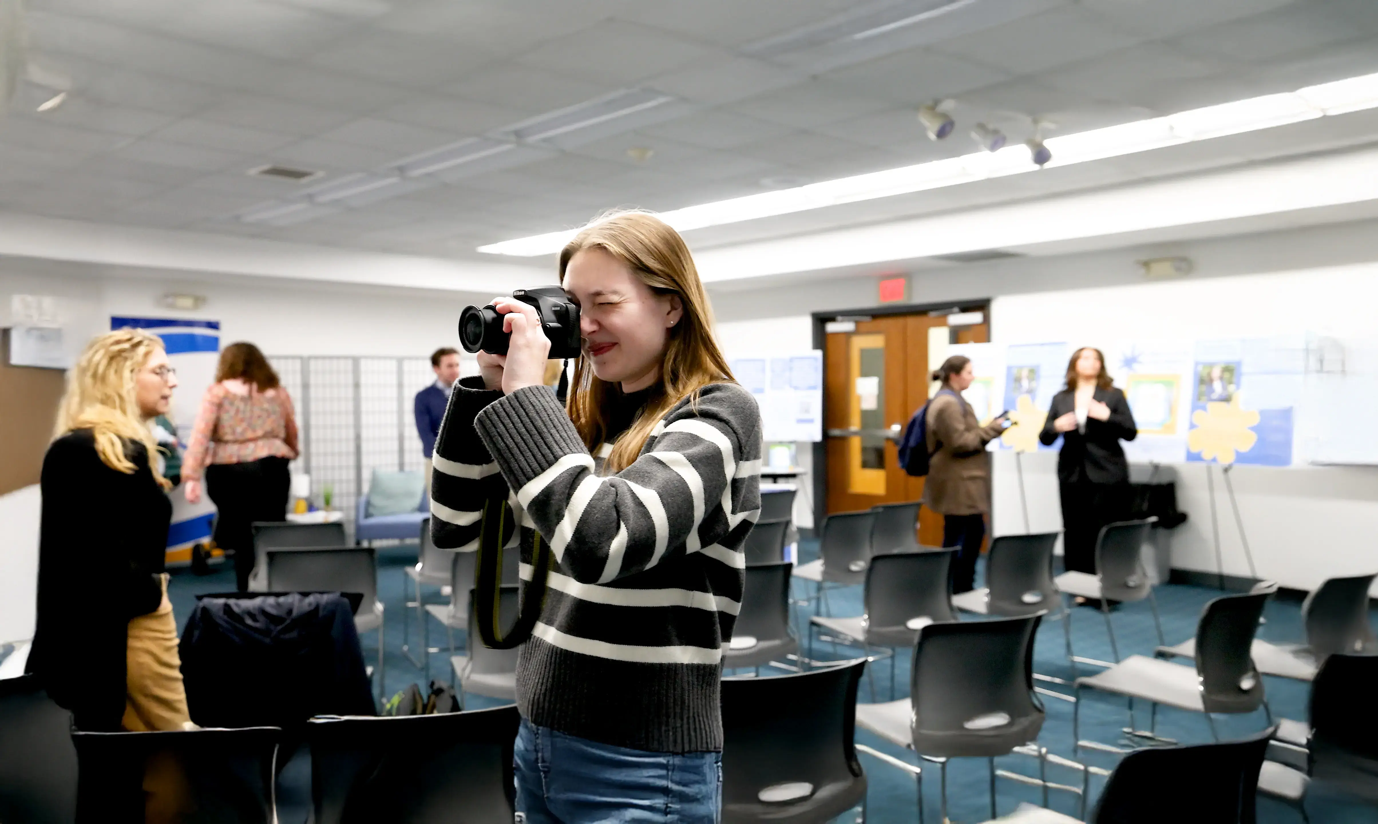 A student takes a photo with a DSLR camera at an indoor event with posters and people in the background.