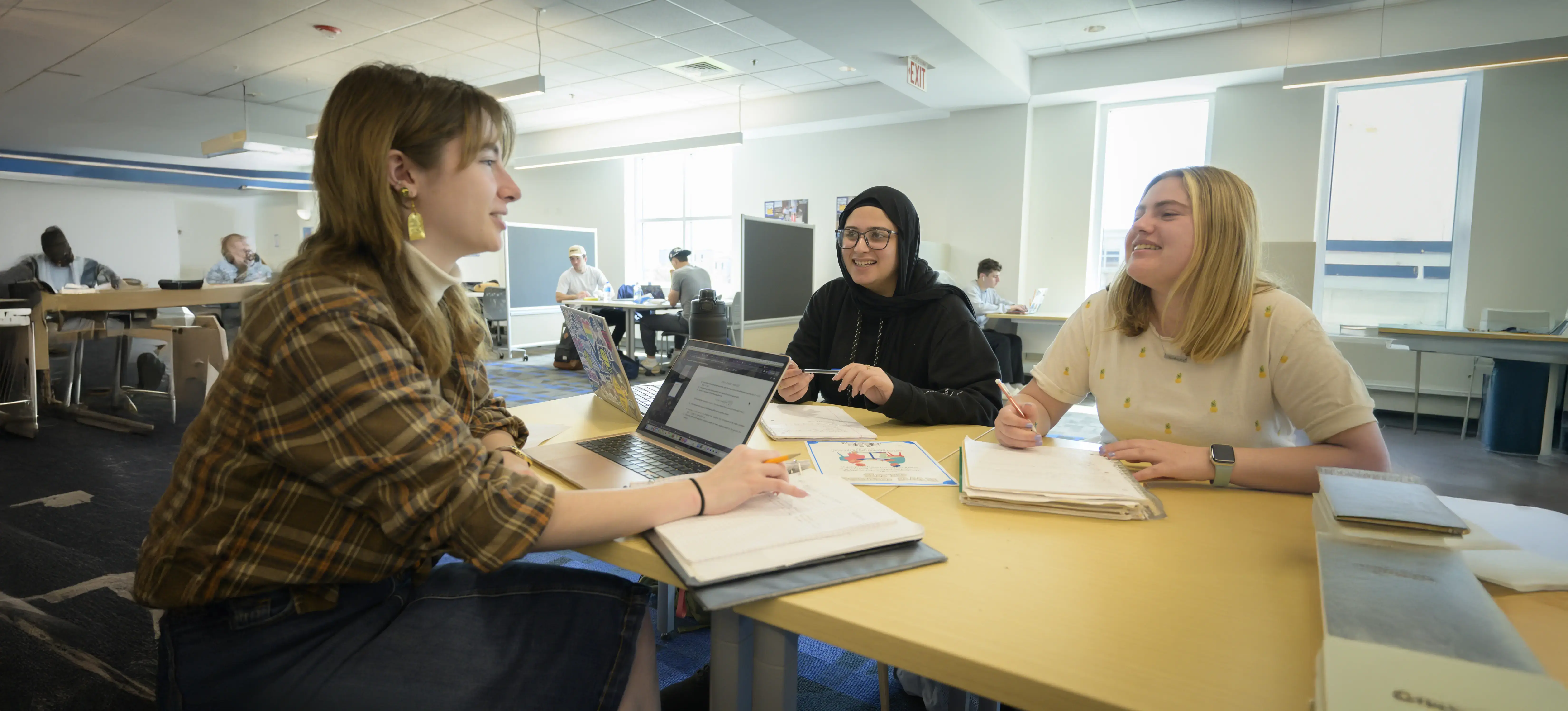Three students have a conversation around a table with books and laptops open in a library setting.