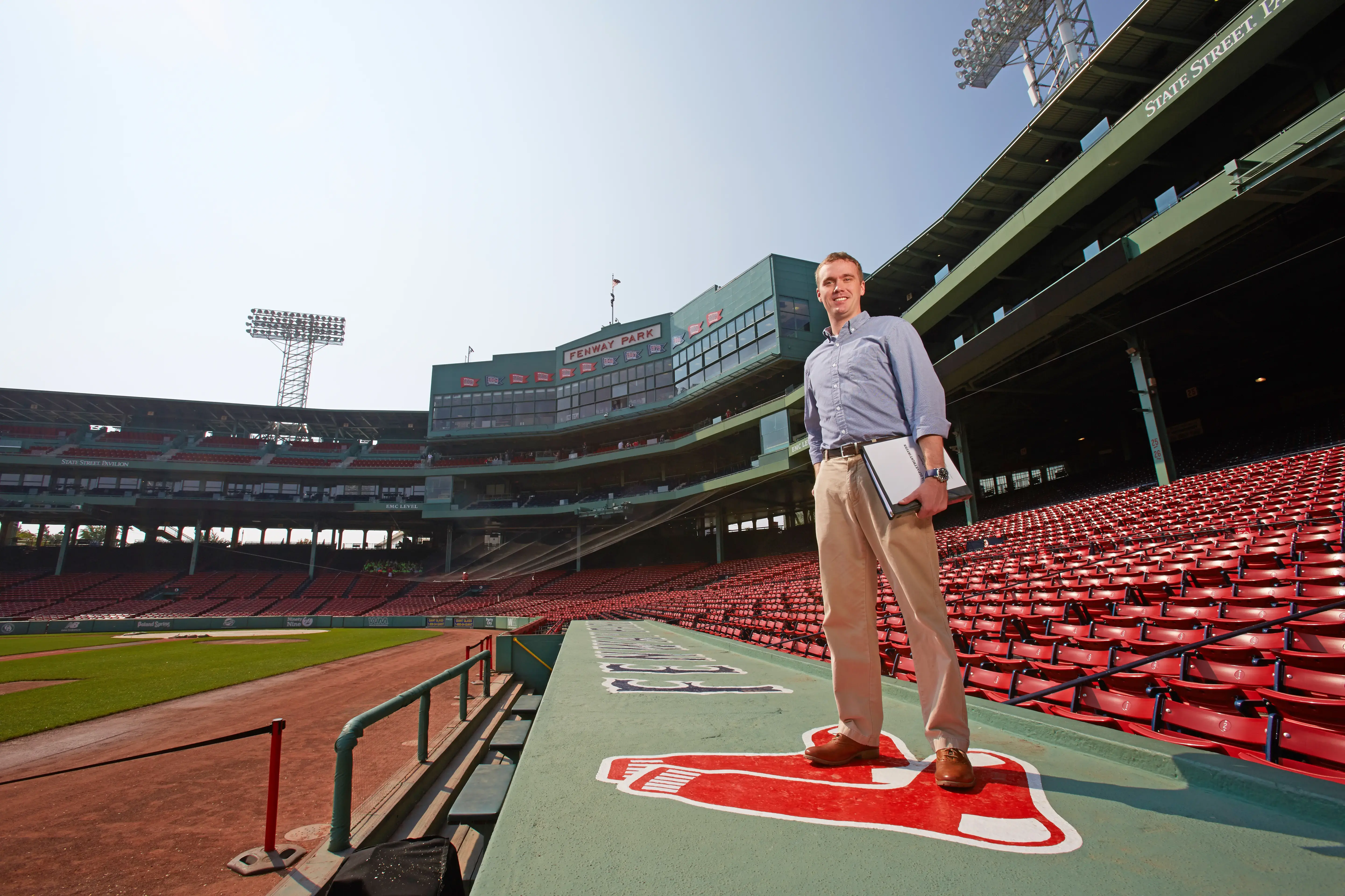 Man holding laptop stands on dugout roof at empty Fenway Park, red seats and scoreboards behind him.