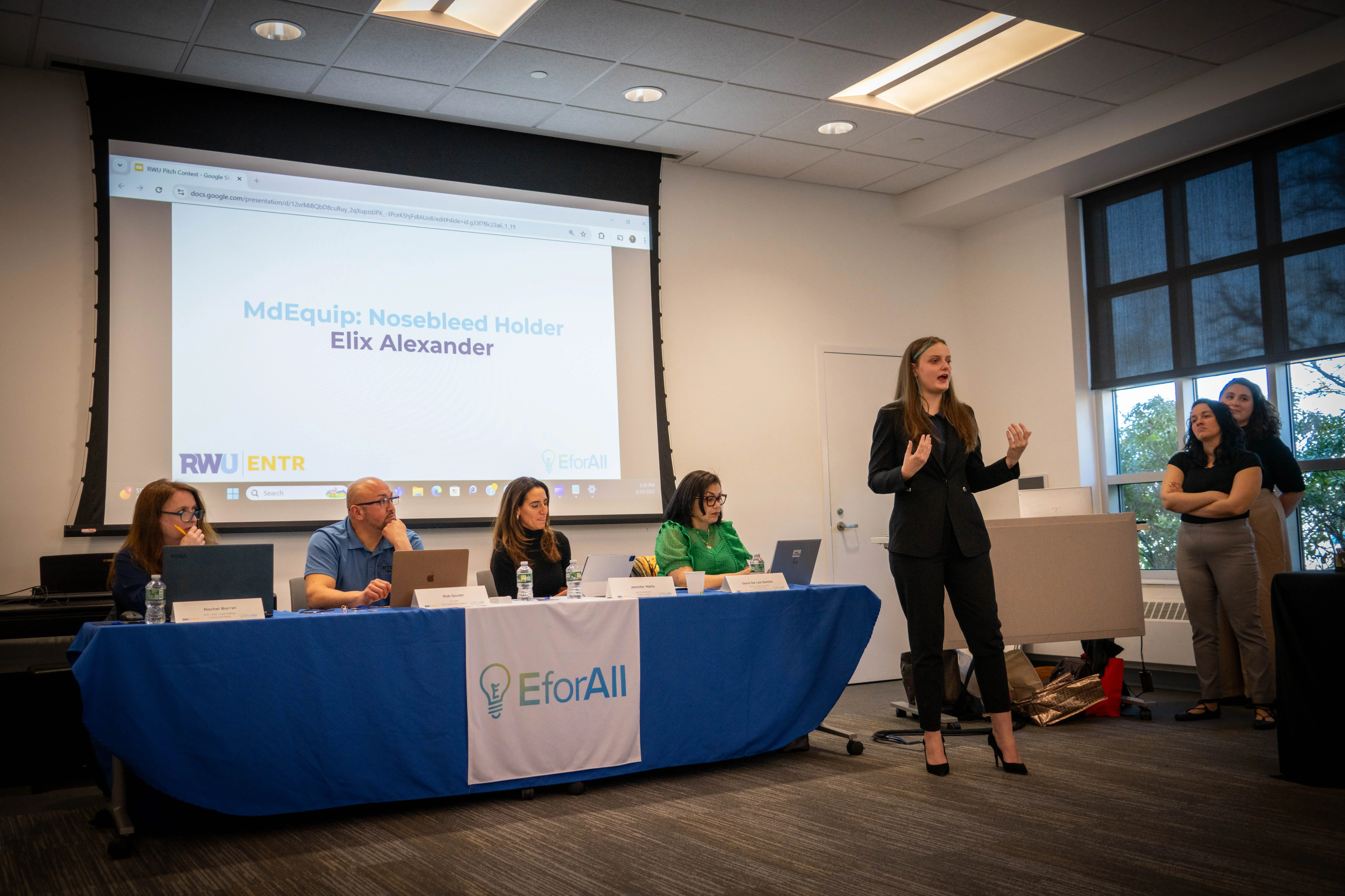 A student pitches product to panel of judges during business competition, with presentation slide behind her.