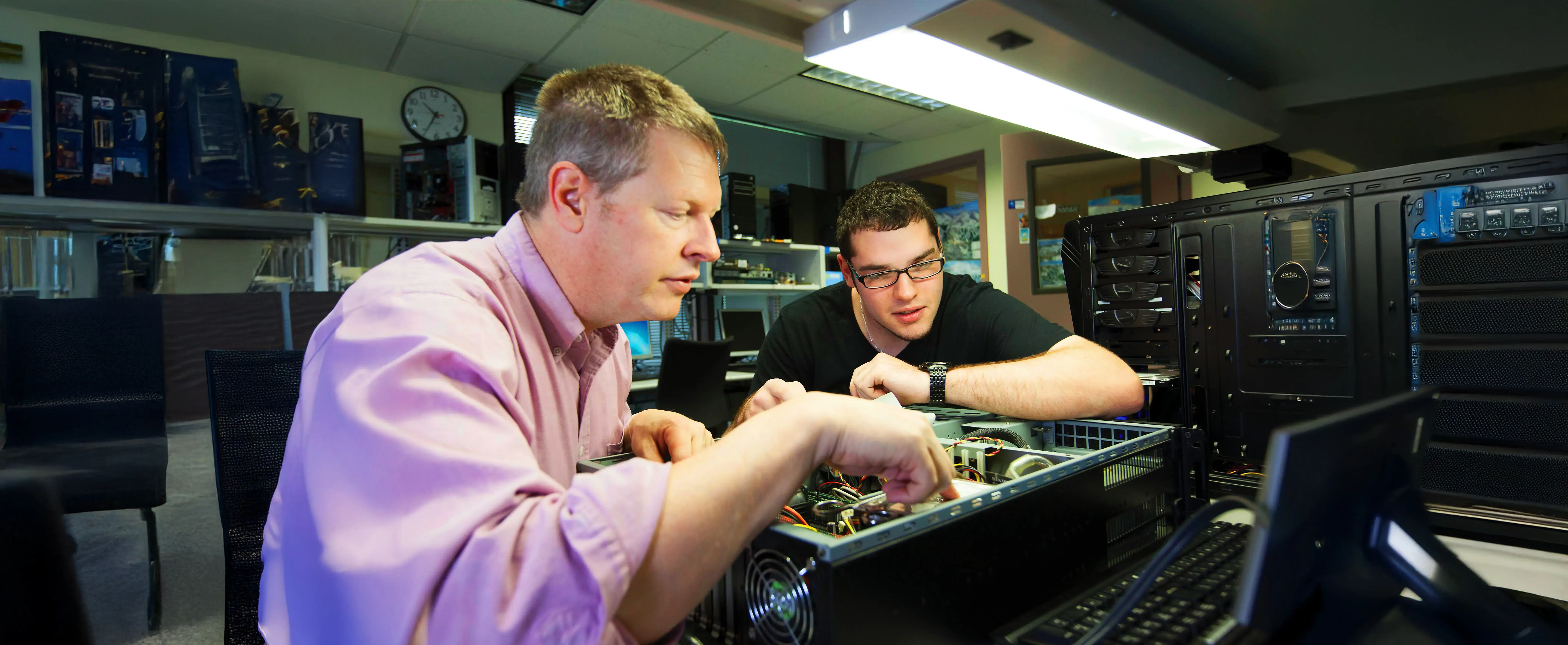 A professor and student examine open computer hardware together in a tech lab with components and monitors in the background.