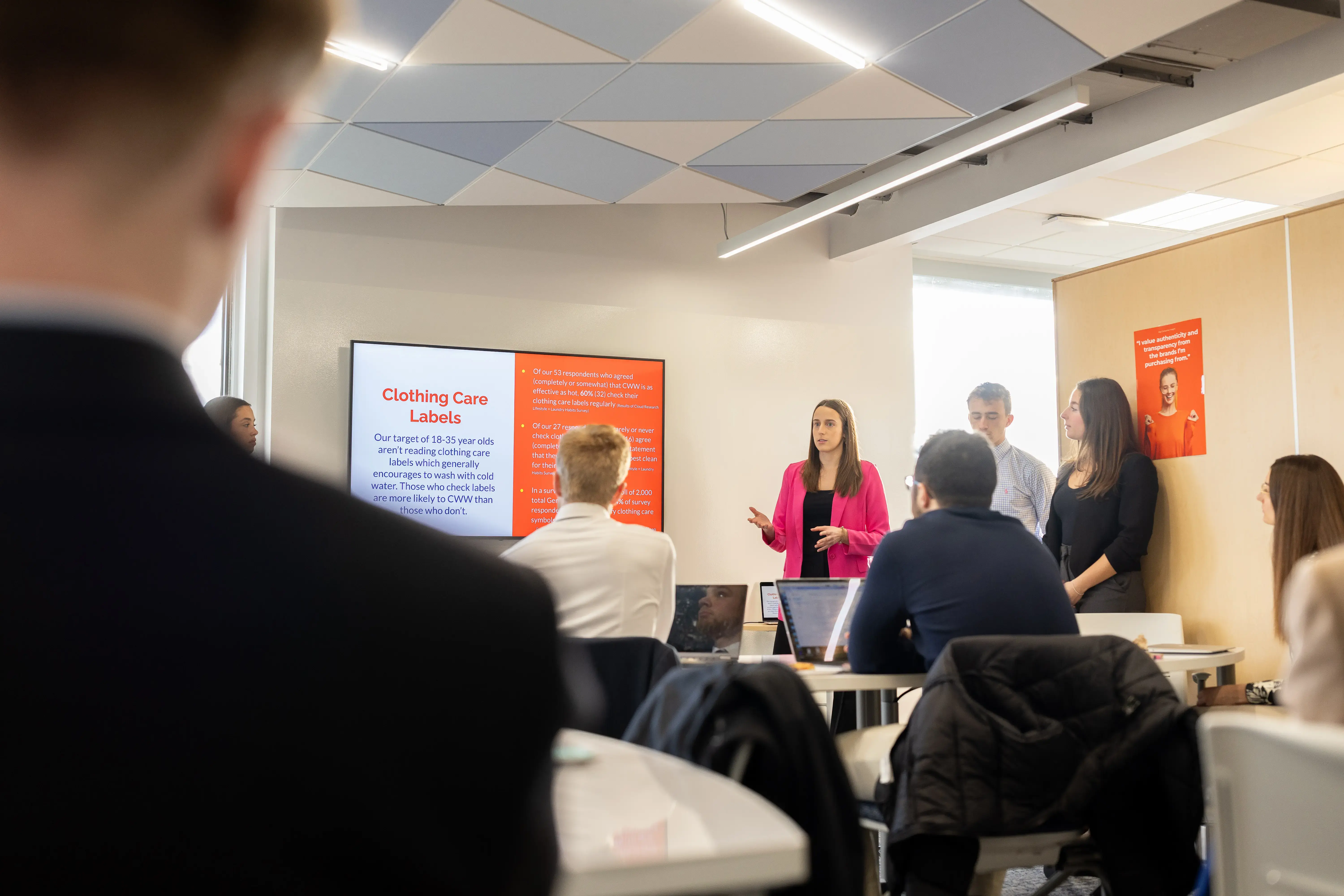 Student presents marketing project on clothing care labels to class, with team and slide display in background.