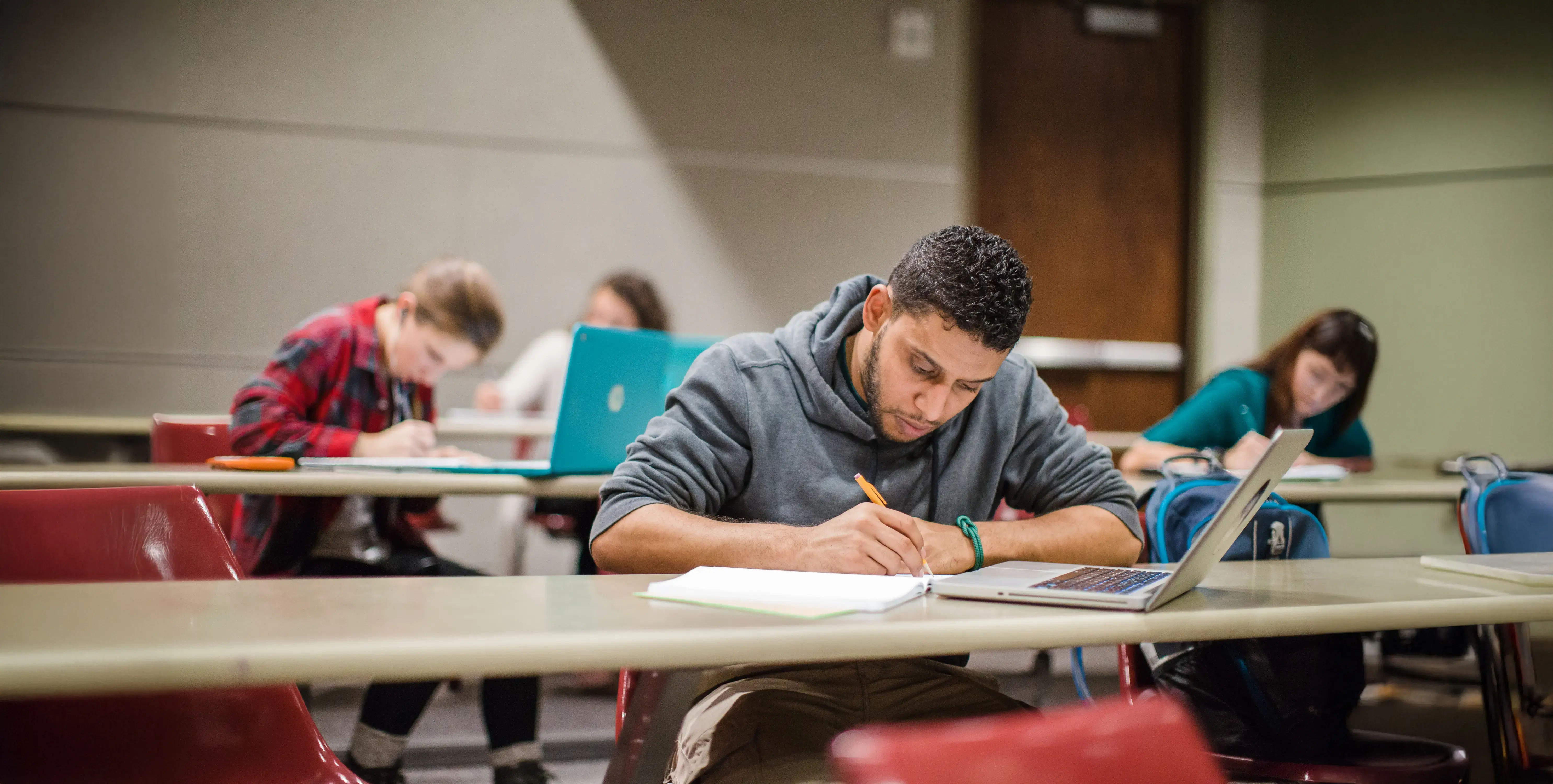 Students sit spaced out in a classroom, focused on writing in notebooks with laptops open beside them.