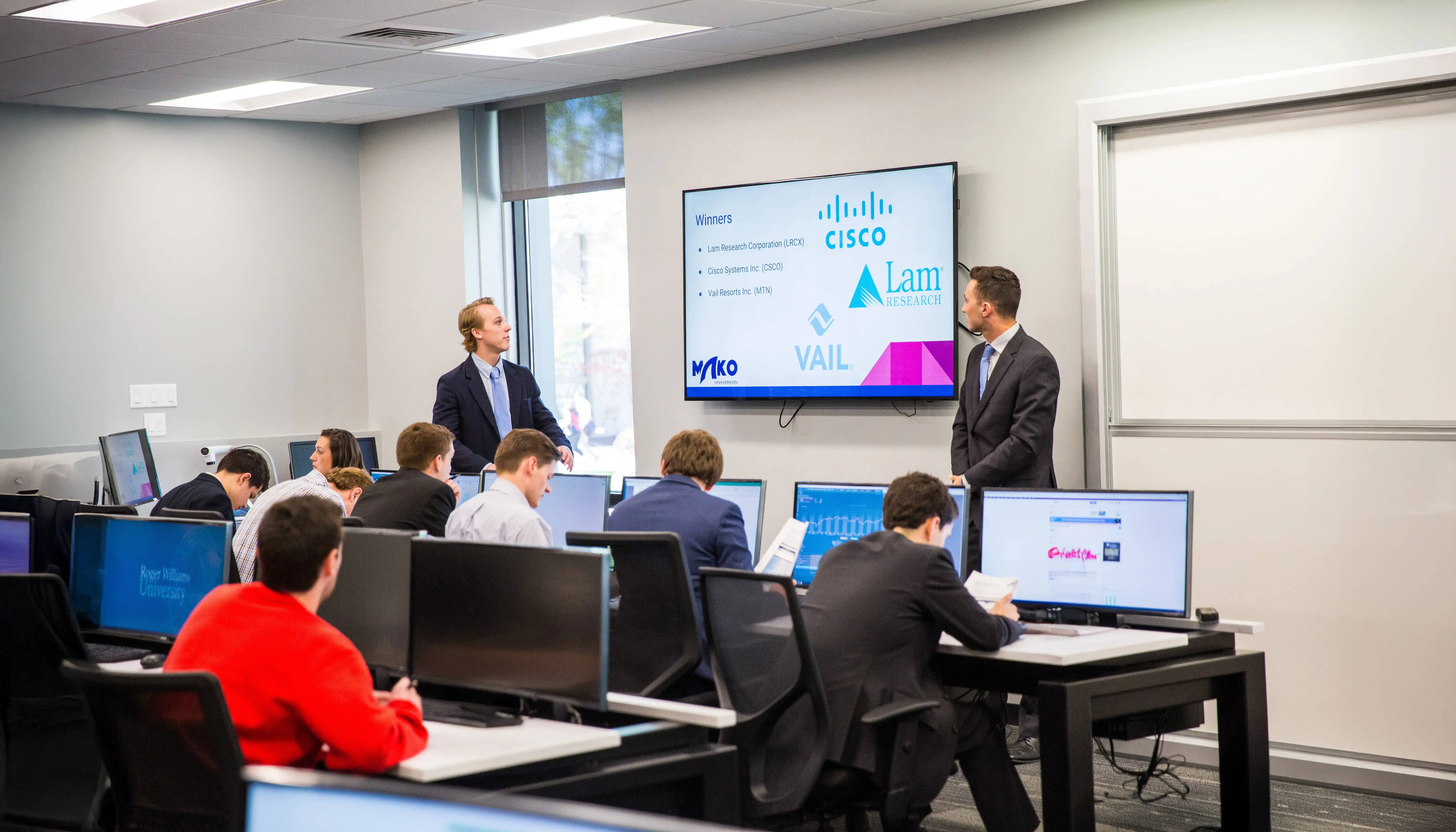 Students in business attire work at computers while two presenters explain less risky investments.