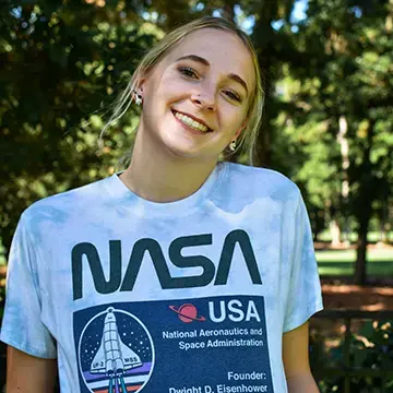 Student in a NASA T-shirt smiles outdoors with trees in the background.