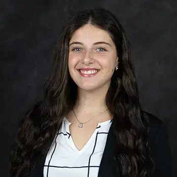 Student in a blazer and patterned blouse poses for a professional portrait against a dark background.