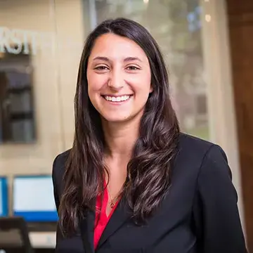 Student in a black blazer and red blouse smiles in a professional setting with glass walls in the background.