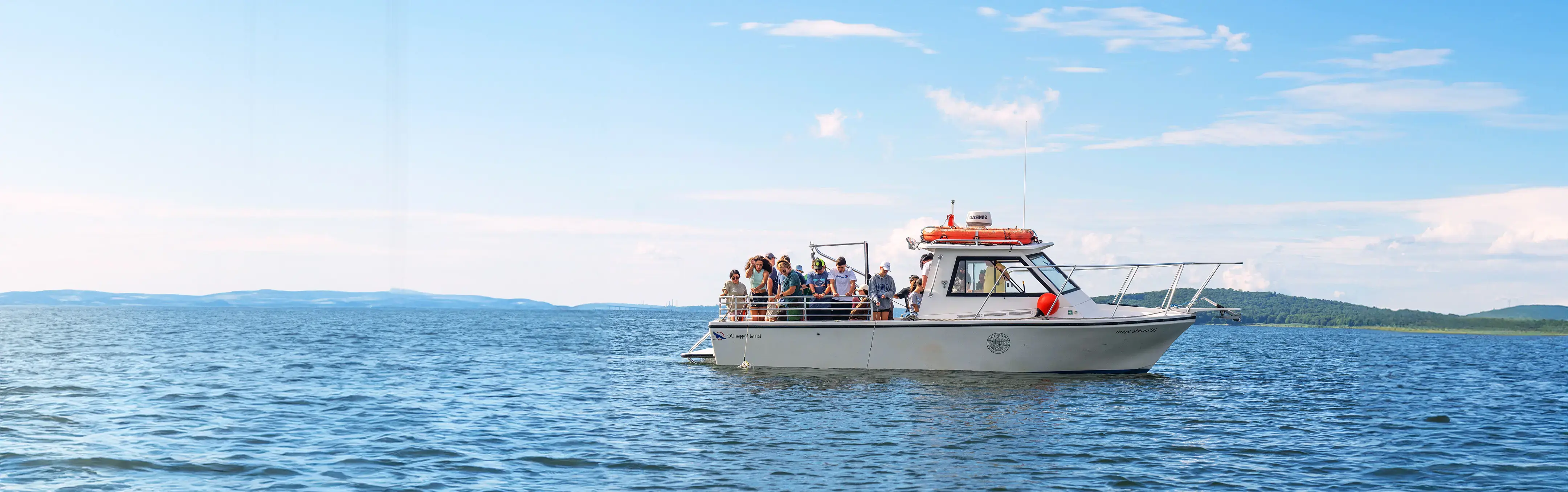 A boat with people on board floats on a calm blue sea under a partly cloudy sky with distant hills visible.