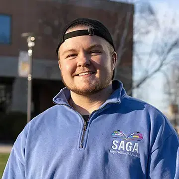 Student wearing a blue SAGA sweatshirt smiles outdoors on the Roger Williams University campus.