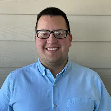 Student in glasses and a light blue button-down shirt smiles in front of a neutral background.