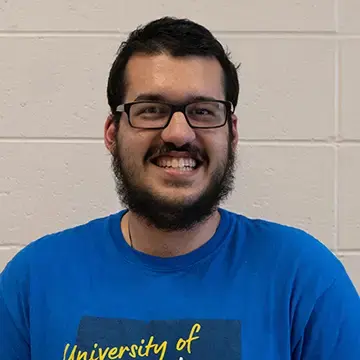 Student in glasses and a blue T-shirt smiles in front of a brick wall.
