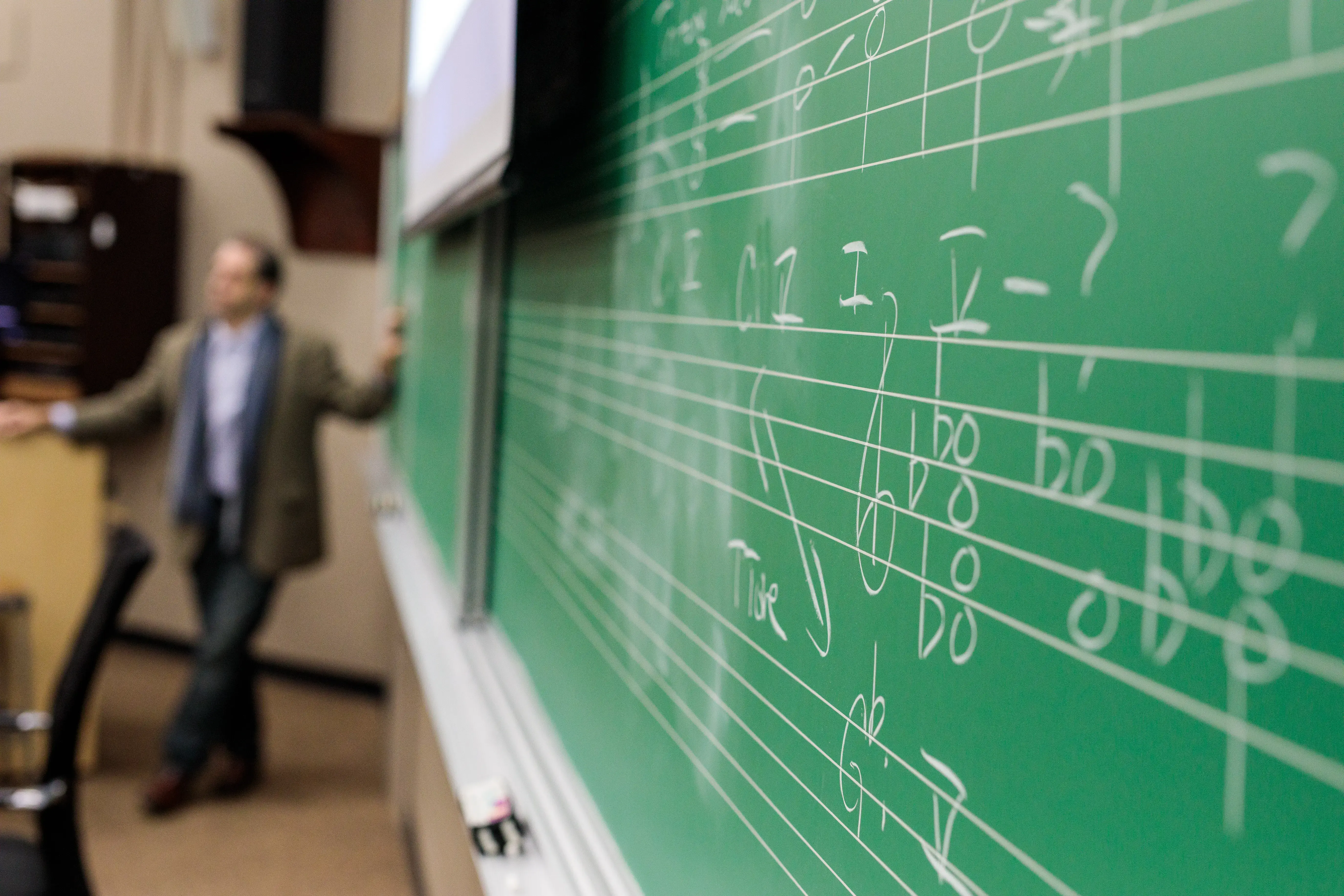 Teacher stands near podium while music notes and chords fill chalkboard in foreground.