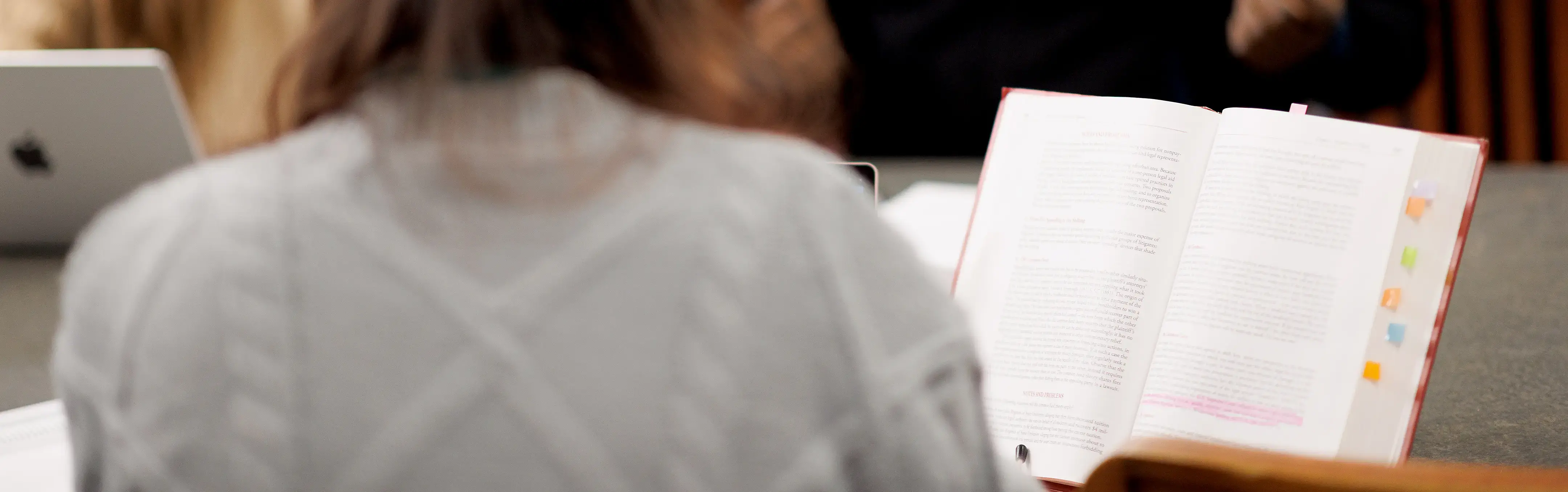 Students gather around a table, discussing with laptops and notes, as a textbook with tabs sits open in the foreground.
