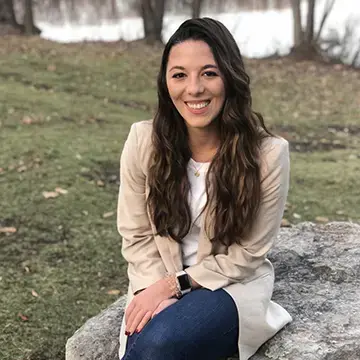 Student in a beige blazer and jeans sits on a rock outdoors near the water.
