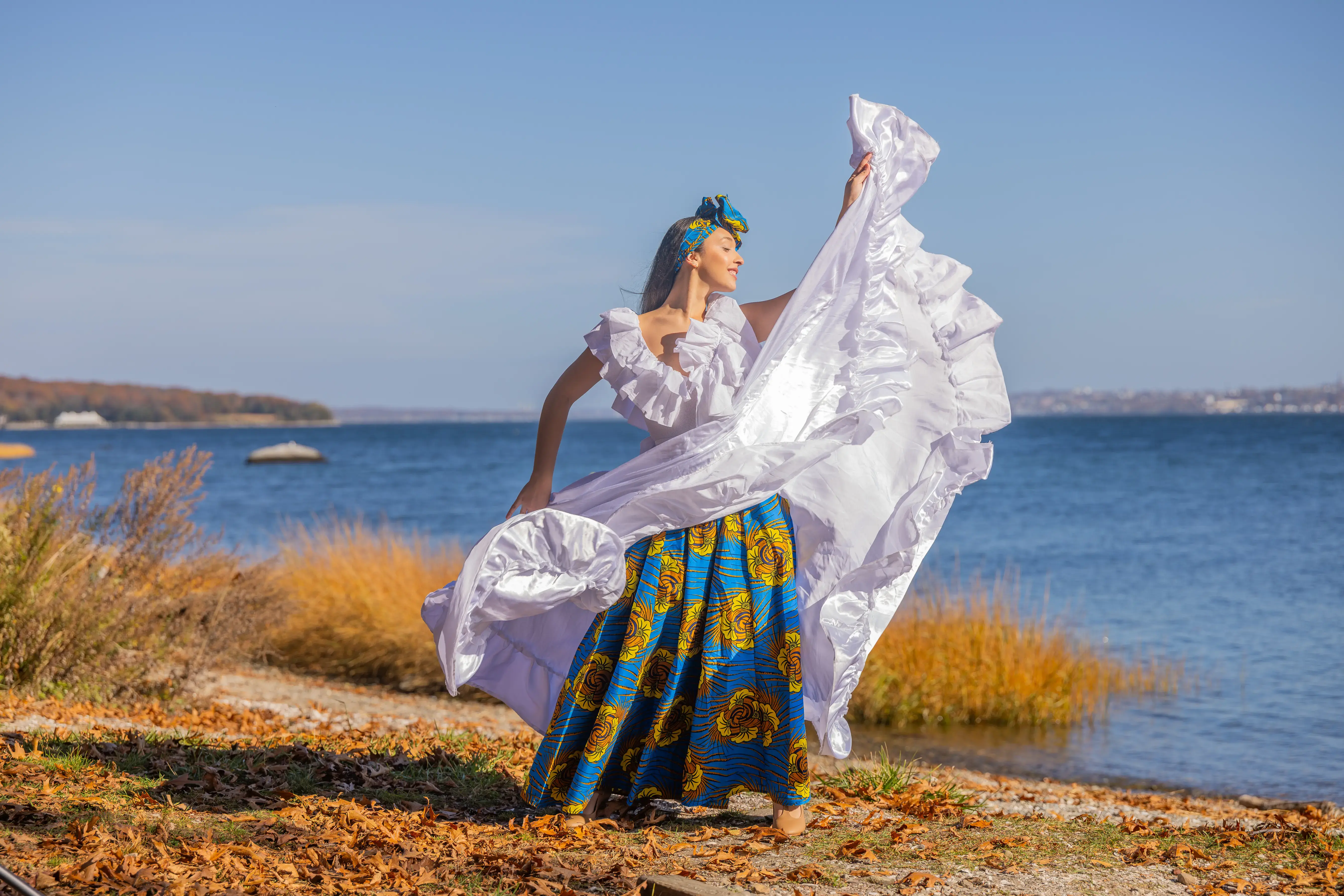 A woman dances in vibrant traditional dress by a lakeside with fall foliage and clear blue sky in the background.
