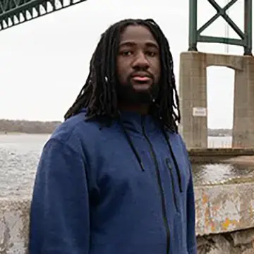 Student in a blue sweatshirt stands outdoors near the Mount Hope Bridge.