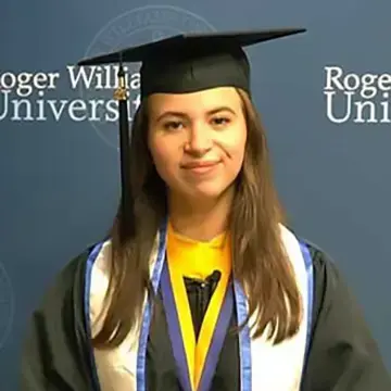 Roger Williams University graduate in cap and gown poses in front of an RWU backdrop.