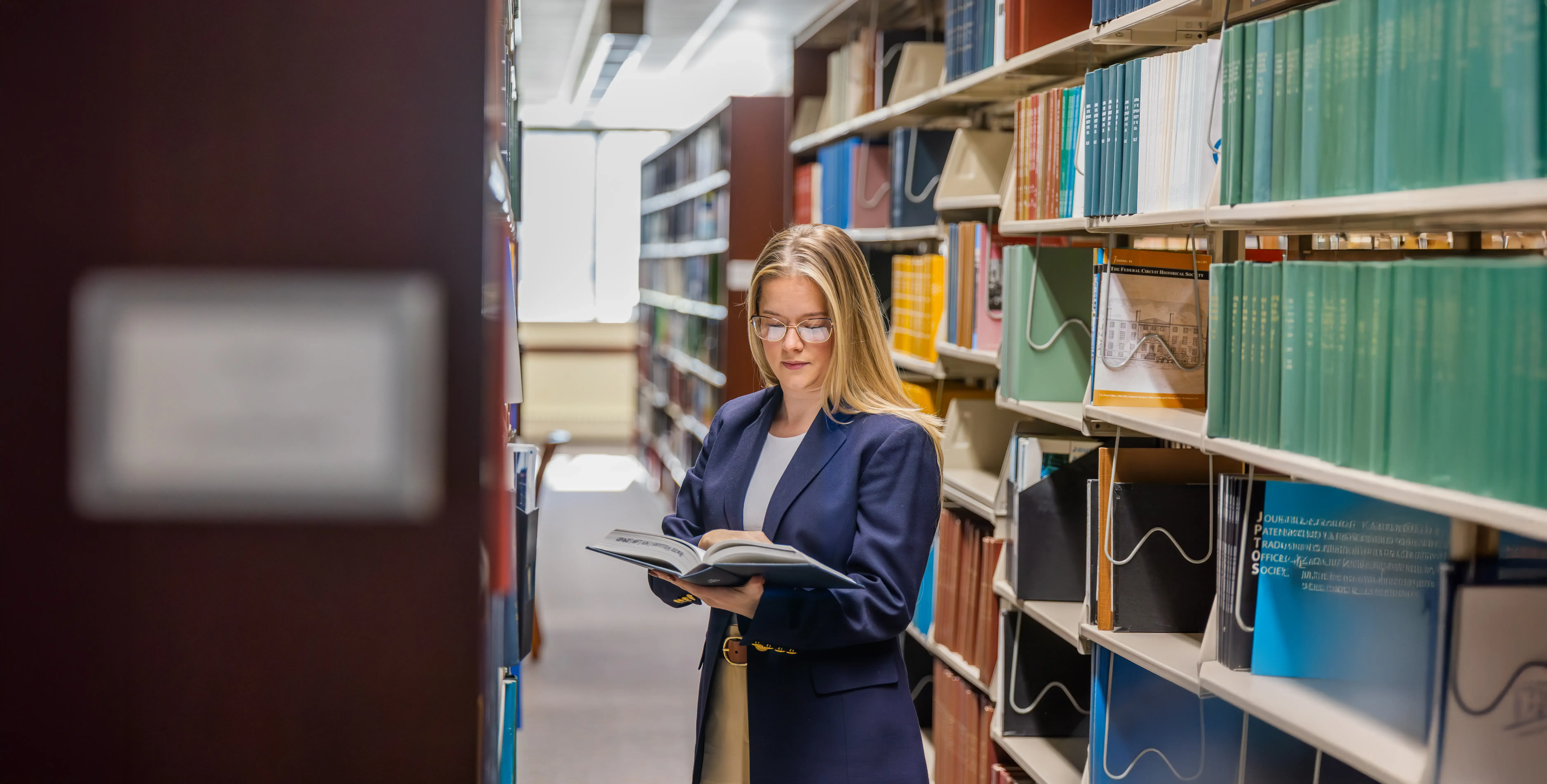 A Law student stands between library shelves reading a book, surrounded by colorful volumes and bright natural light from nearby windows.