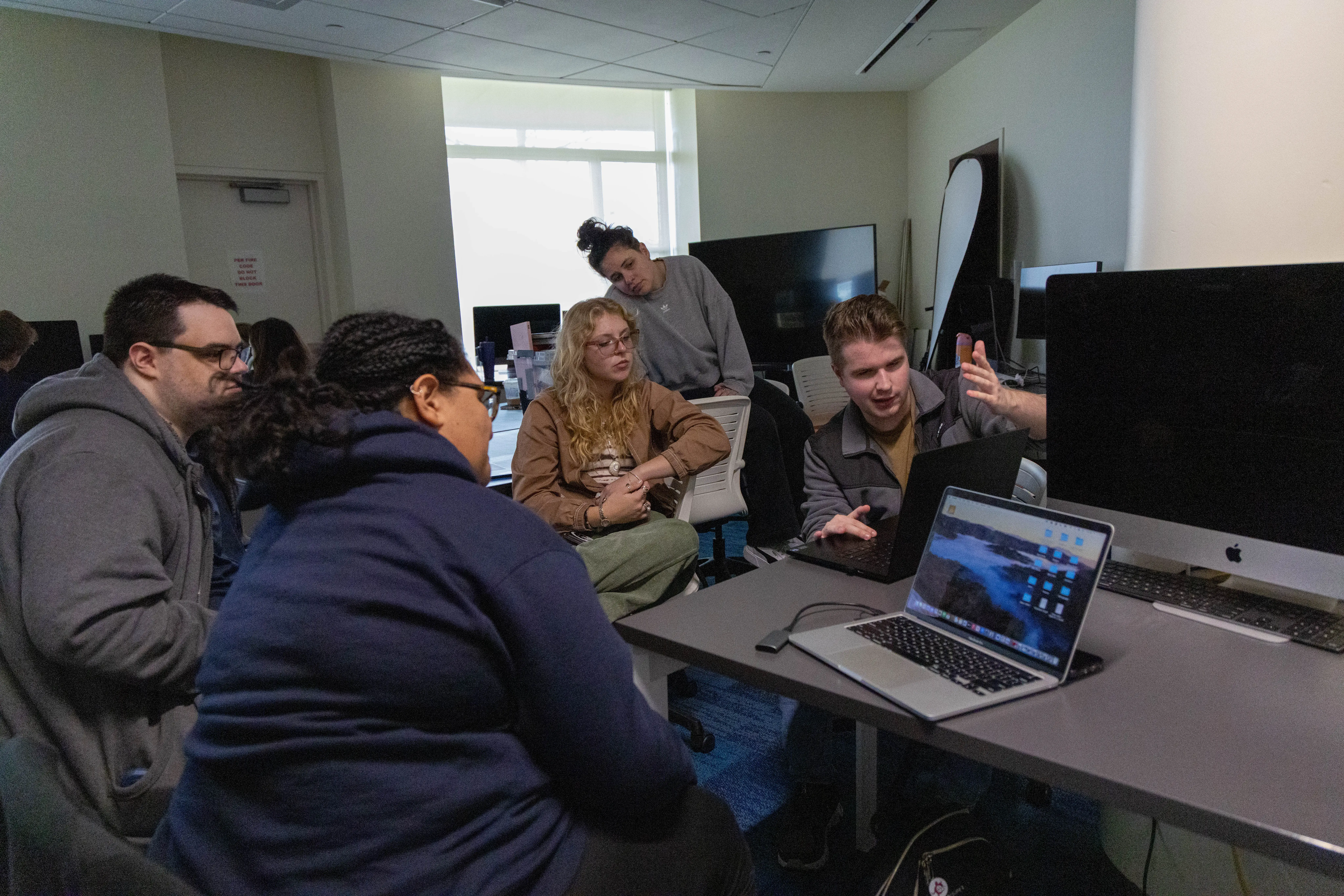 Students gather around a laptop in a dim computer lab, discussing a project surrounded by desktop monitors and iMacs.