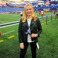Alumna stands on a football field holding a microphone during a sports broadcast.