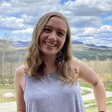 Student in a tank top smiles outdoors with mountains and blue sky in the background.