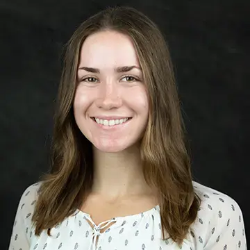 Student in a white patterned blouse smiles in front of a dark background.